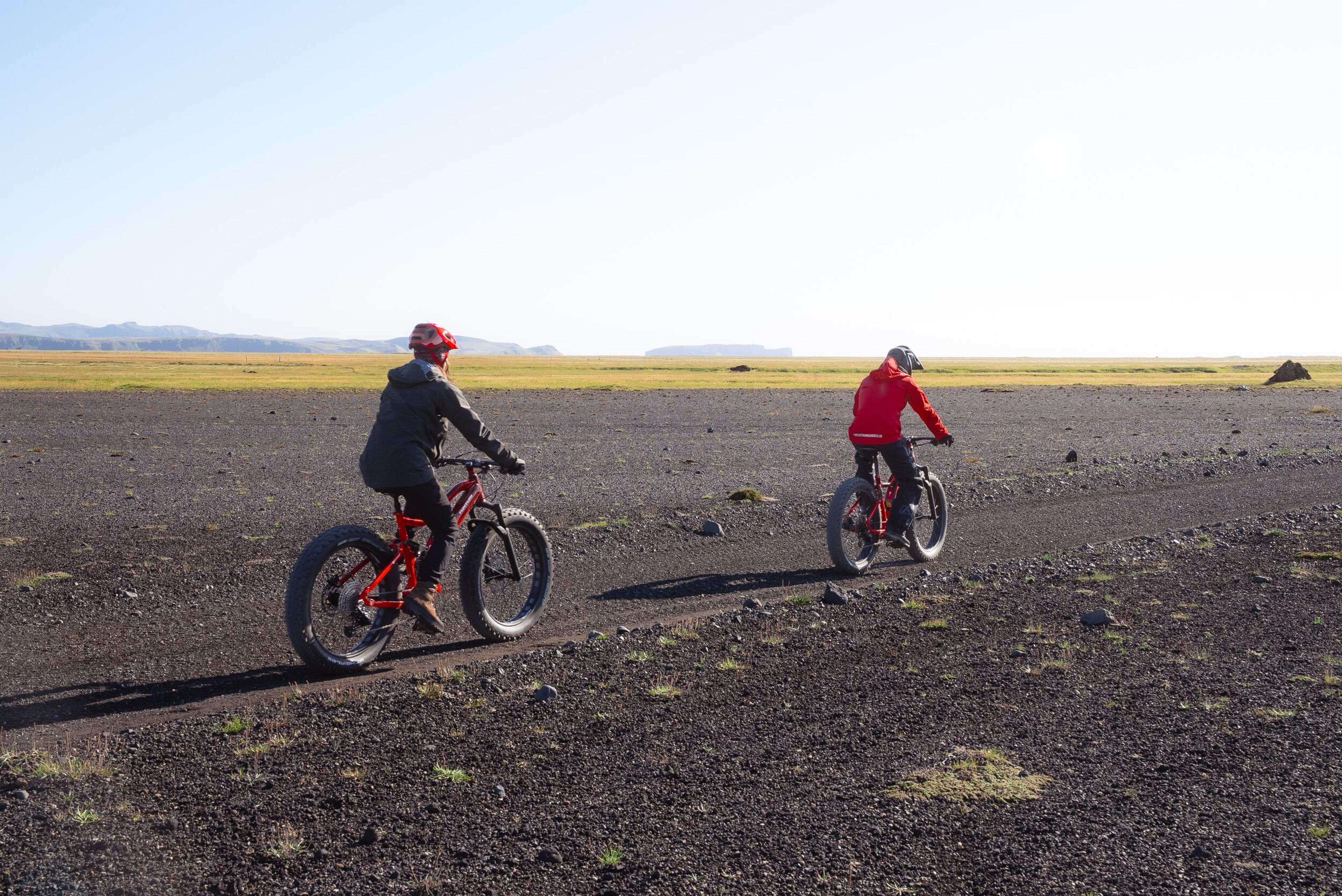 Two people ride fat-tire bikes on a dark, rocky plain with a green field in the distance.