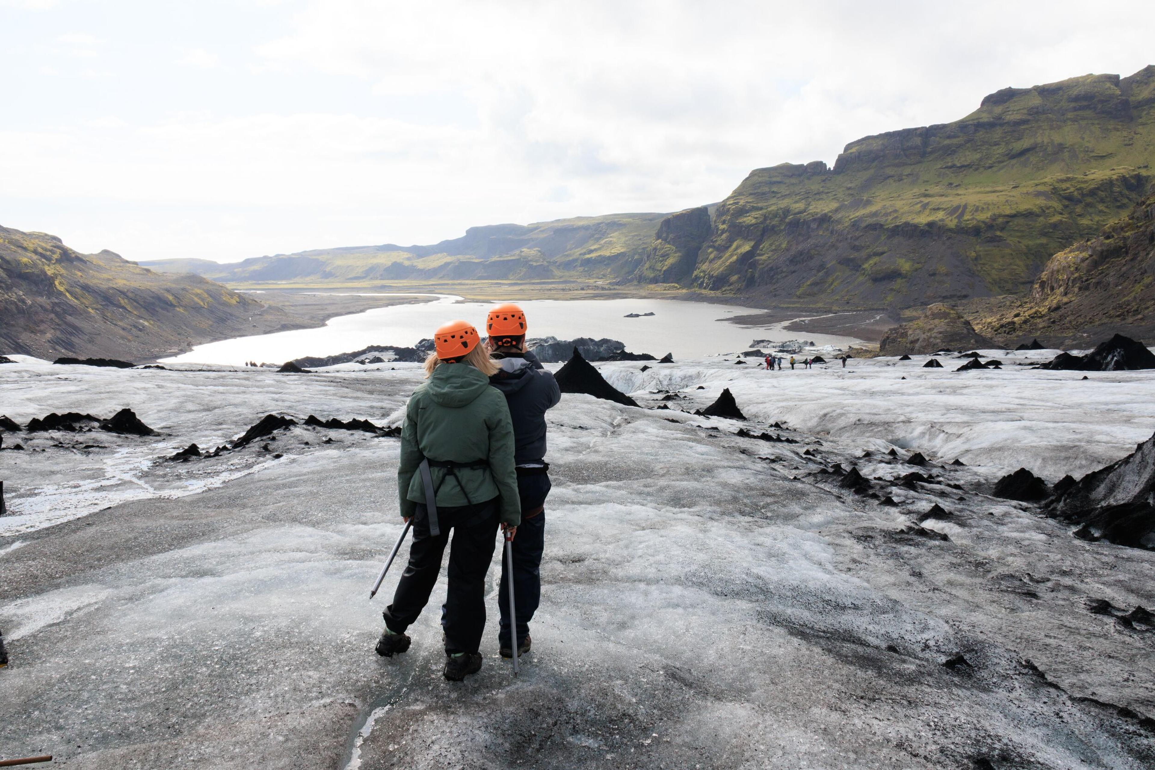 Two people in helmets and harnesses stand on a dark, ash-covered glacier, looking out at a glacial lake and mountains.