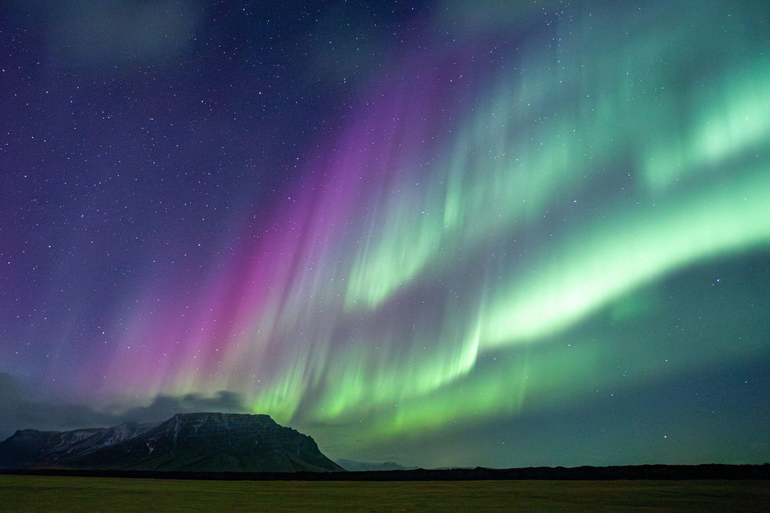 Purple and green aurora borealis lights up a starry night sky above a dark mountain range and field.
