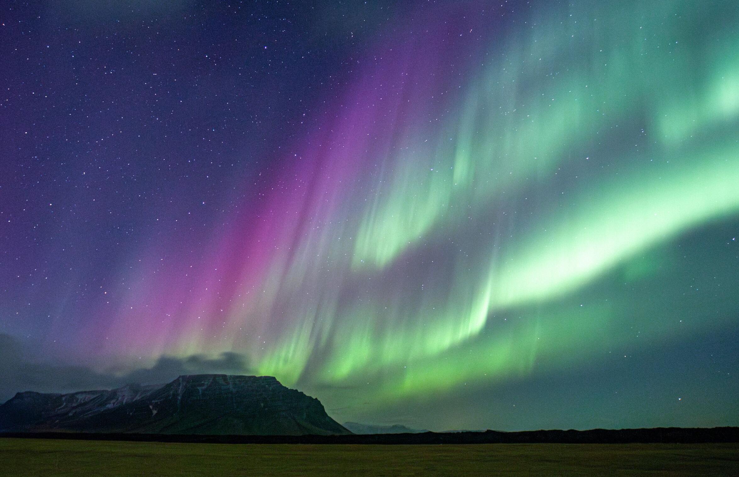 Purple and green aurora borealis lights up a starry night sky above a dark mountain range and field.