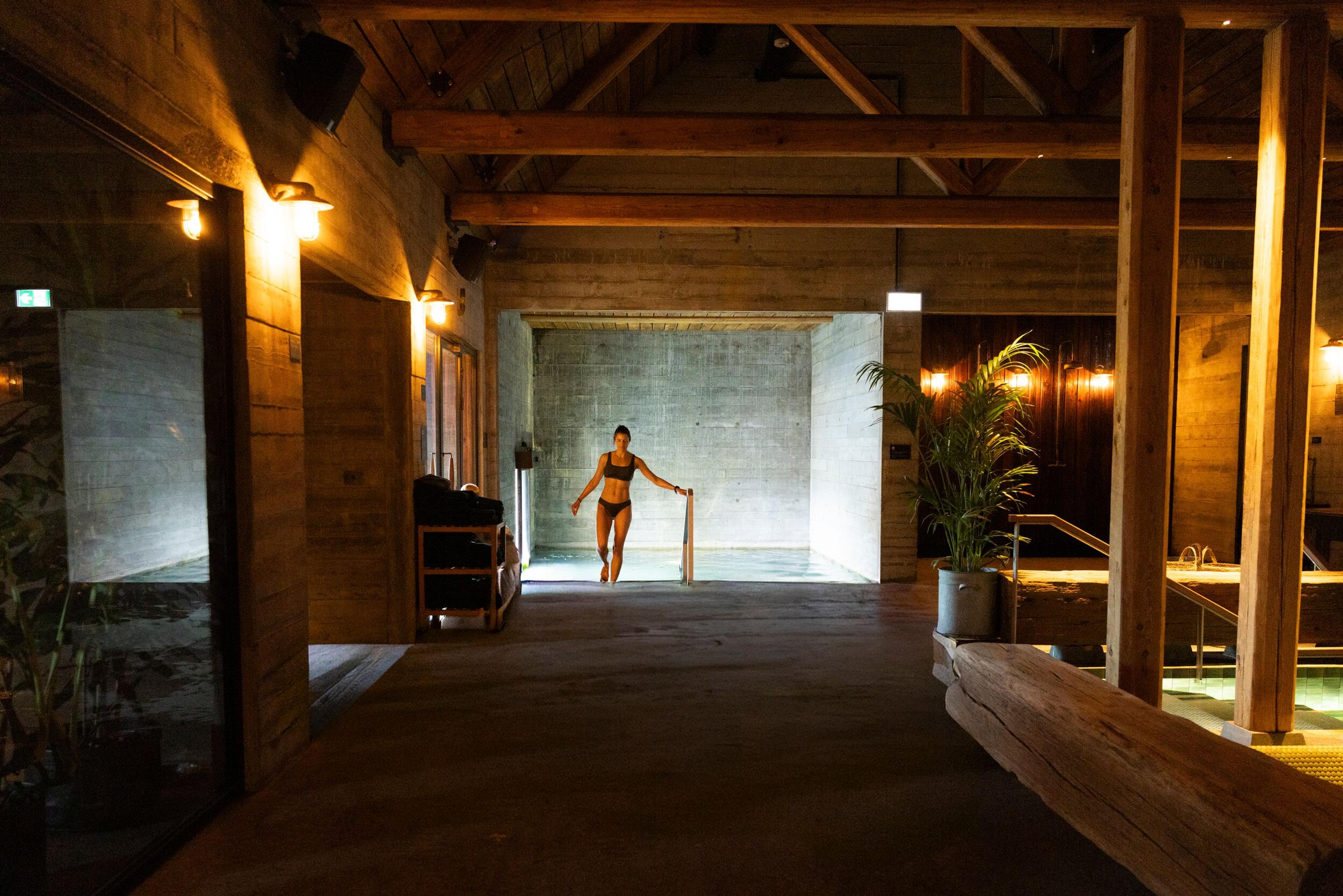A woman in a dark swimsuit steps into a concrete-walled pool under warm lighting and wooden beams.