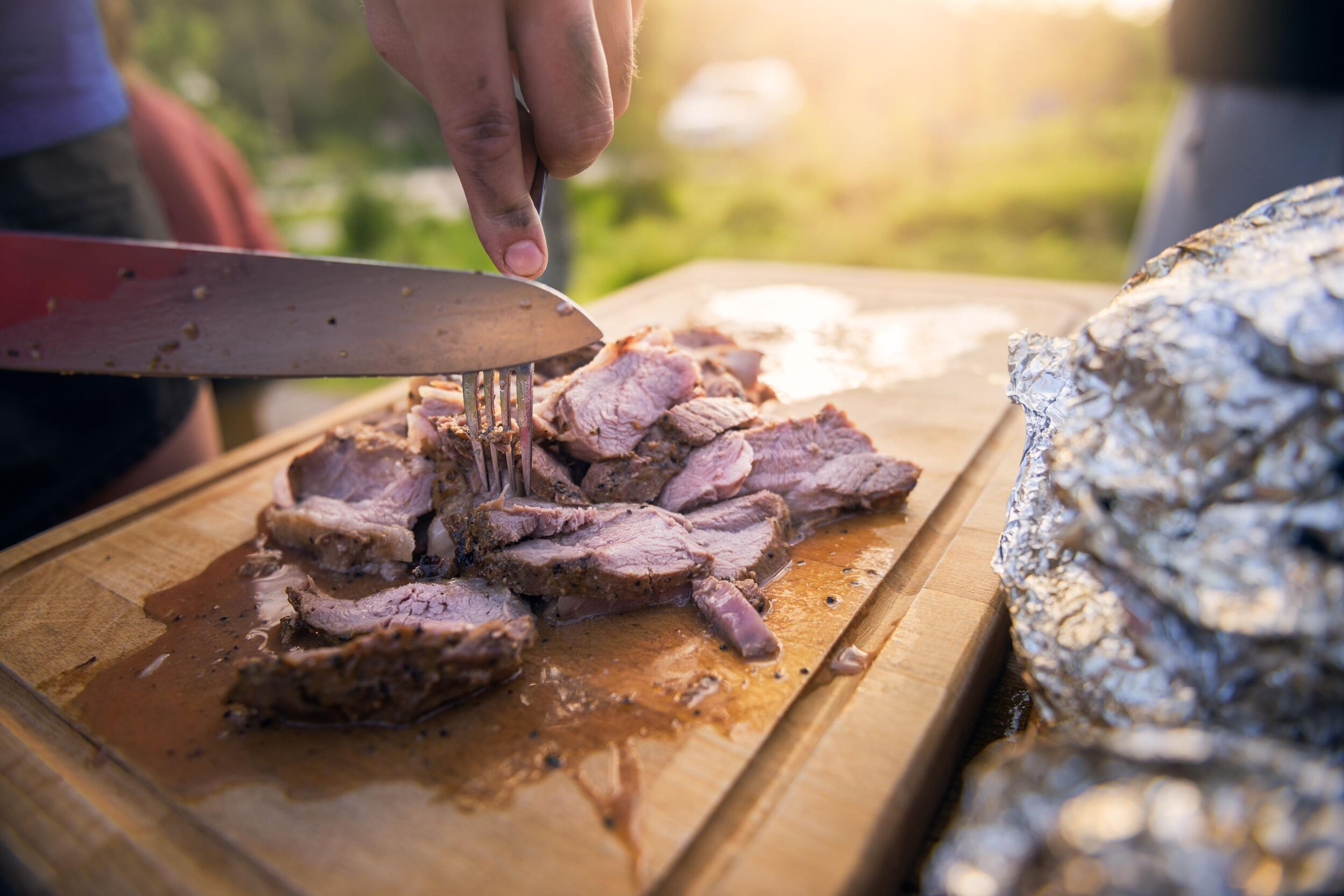A person slices cooked meat on a wooden cutting board outdoors.