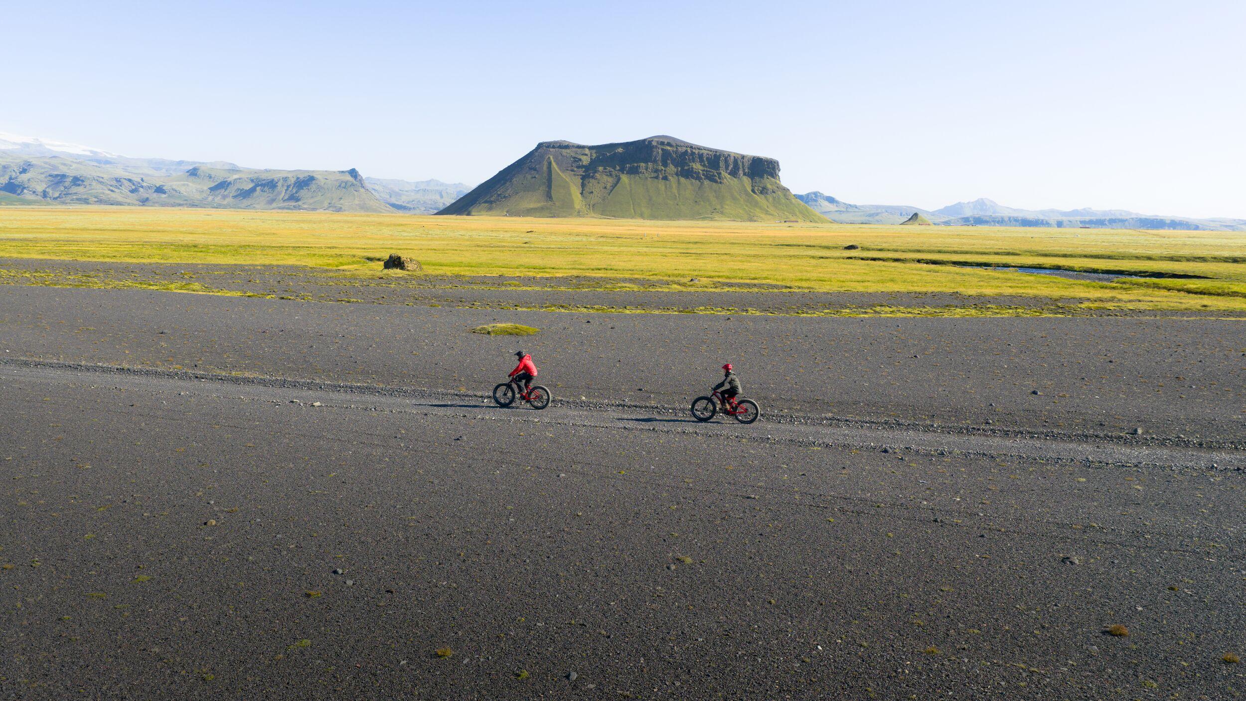 Two people ride fat-tire bikes across a black gravel plain with green fields and flat-topped mountains in the background.