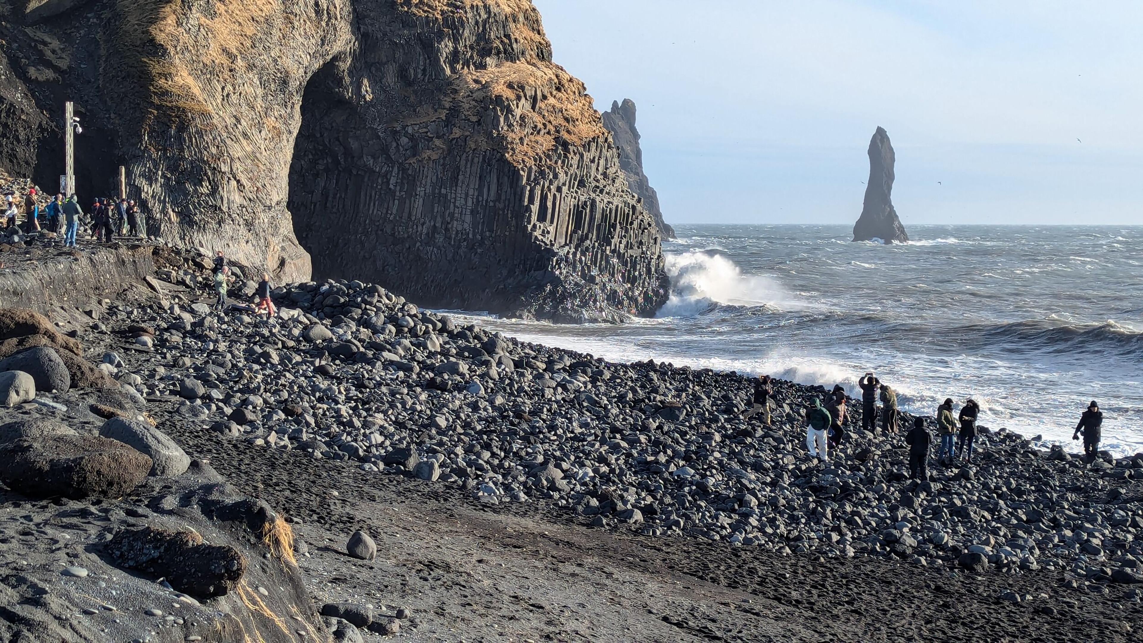 A black pebble beach with a basalt sea cave, tourists, crashing waves, and a tall sea stack in the distance.