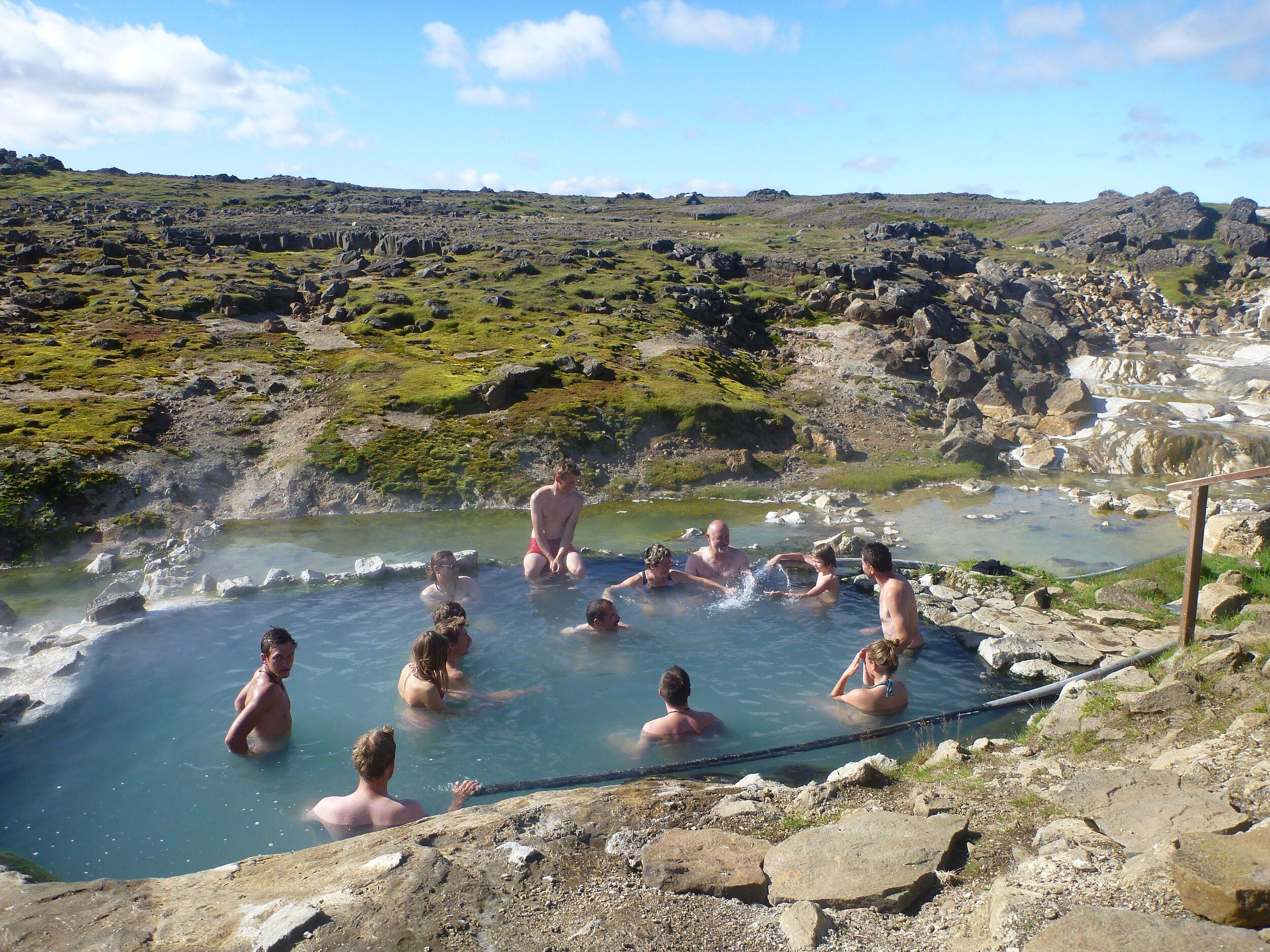 People bathing in a natural, steaming hot spring surrounded by rocky, green hills.