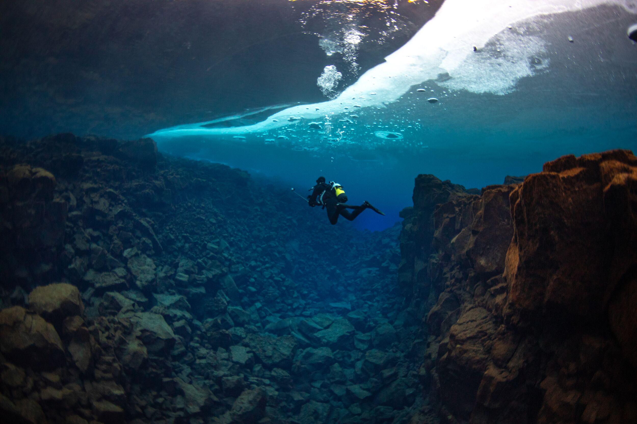 A diver in a clear blue underwater rift with an ice sheet above.