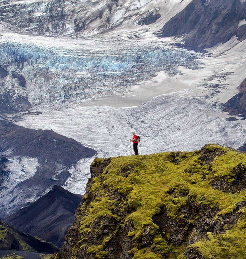 A lone hiker in a red jacket stands on the edge of the cliff, gazing at the icy expanse below. The scene highlights the dramatic landscapes of Iceland, where lush volcanic terrain meets the raw power of glaciers.