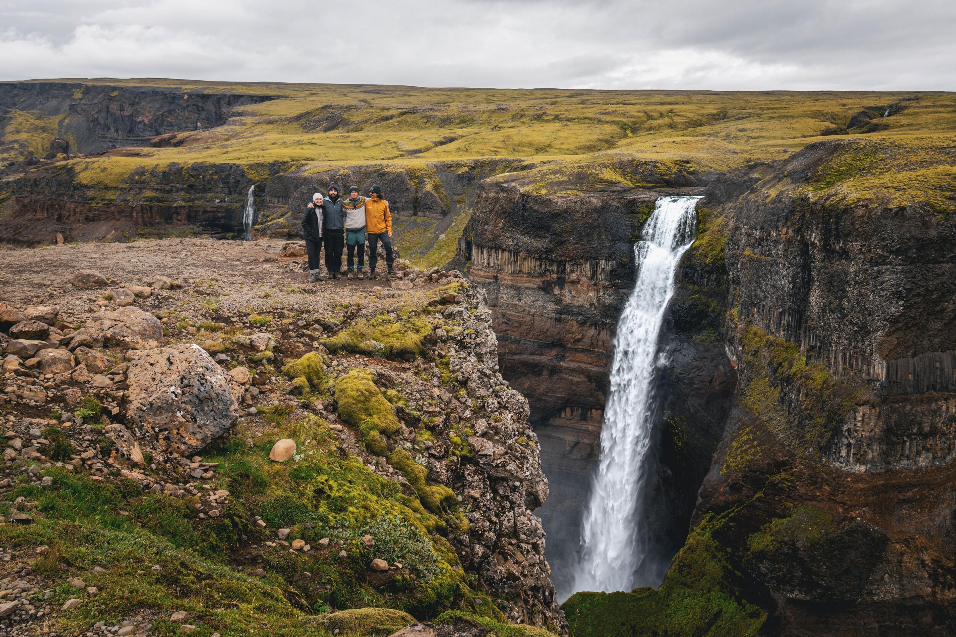 Four people stand on a mossy cliff overlooking a powerful waterfall plunging into a rugged canyon.