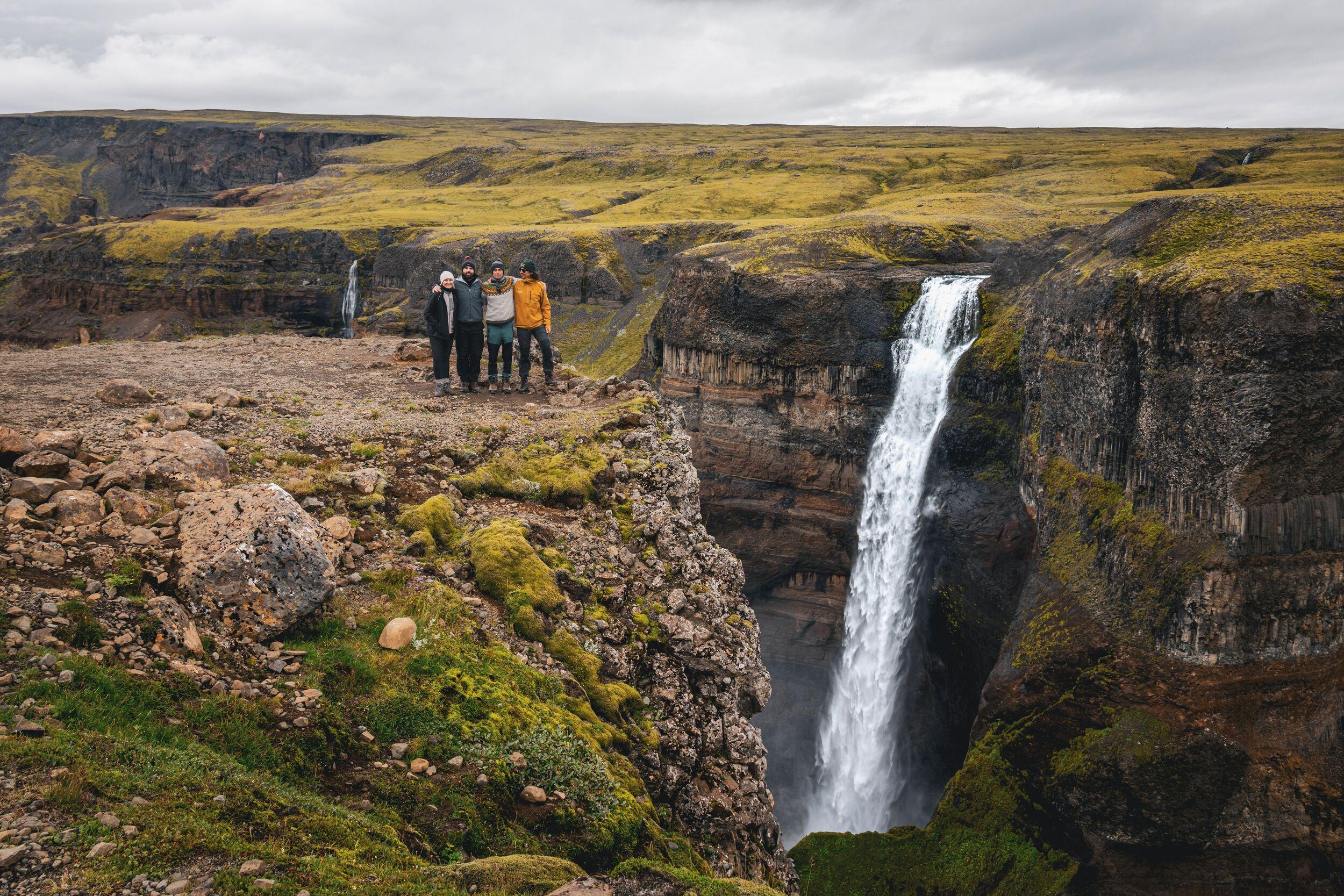 Four people stand on a mossy cliff overlooking a powerful waterfall plunging into a rugged canyon.
