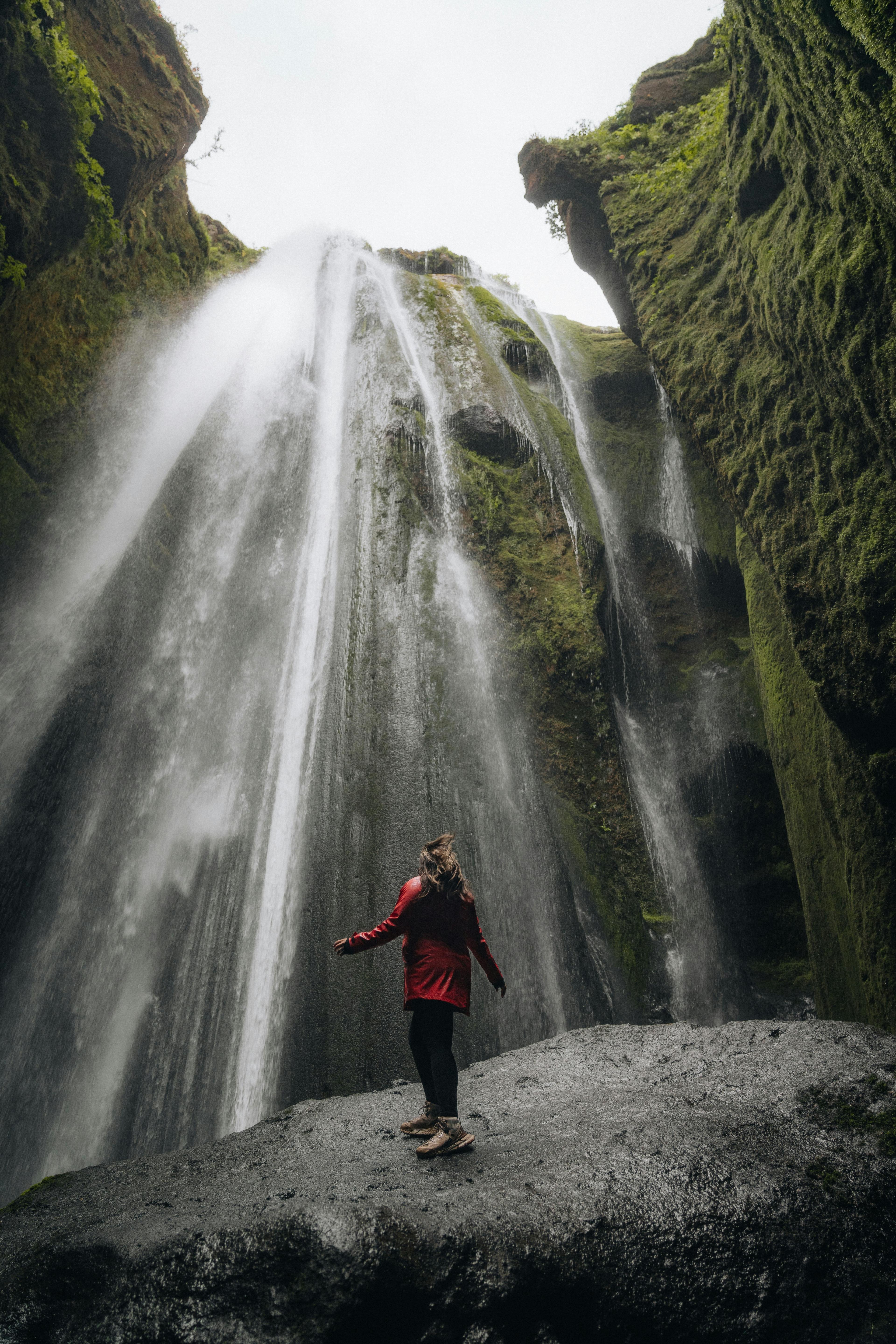 A woman in a red jacket stands at the base of Gljúfrabúi waterfall, surrounded by towering moss-covered cliffs and cascading water.