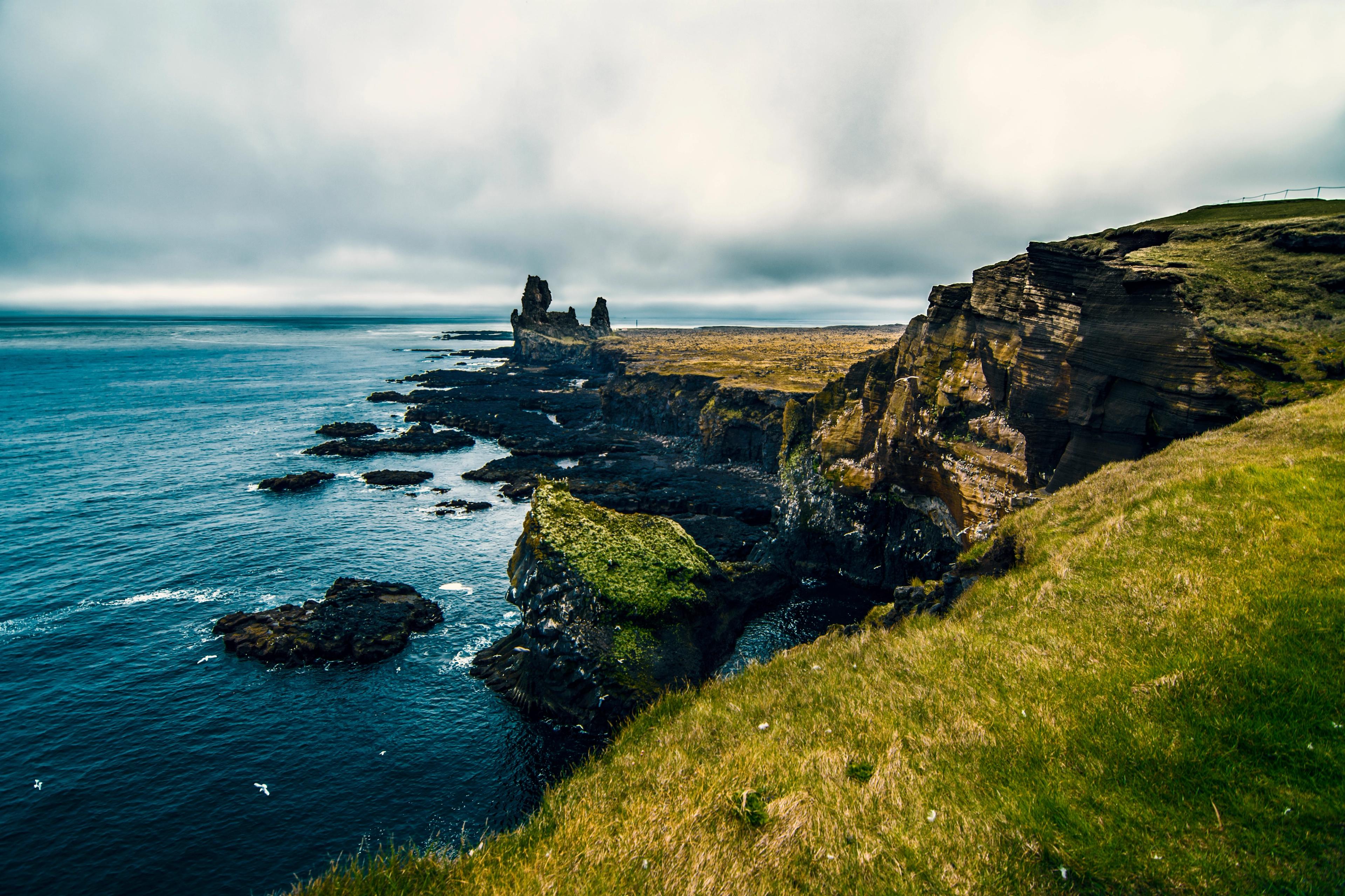 Rugged coastal cliffs with green grass overlooking dark sea stacks and deep blue water under a cloudy sky.