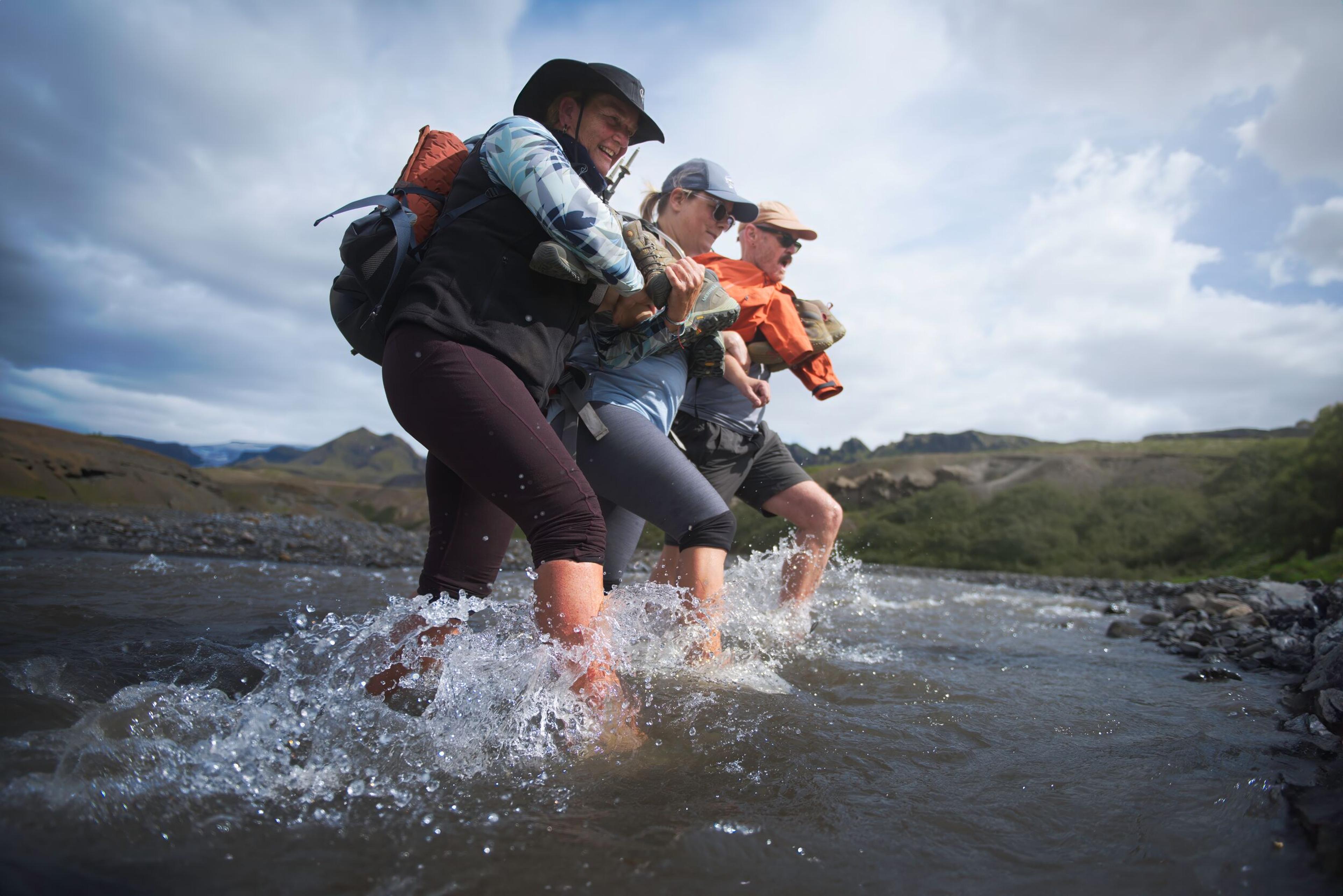 Three hikers carefully crossing a river on foot, holding onto each other for support.