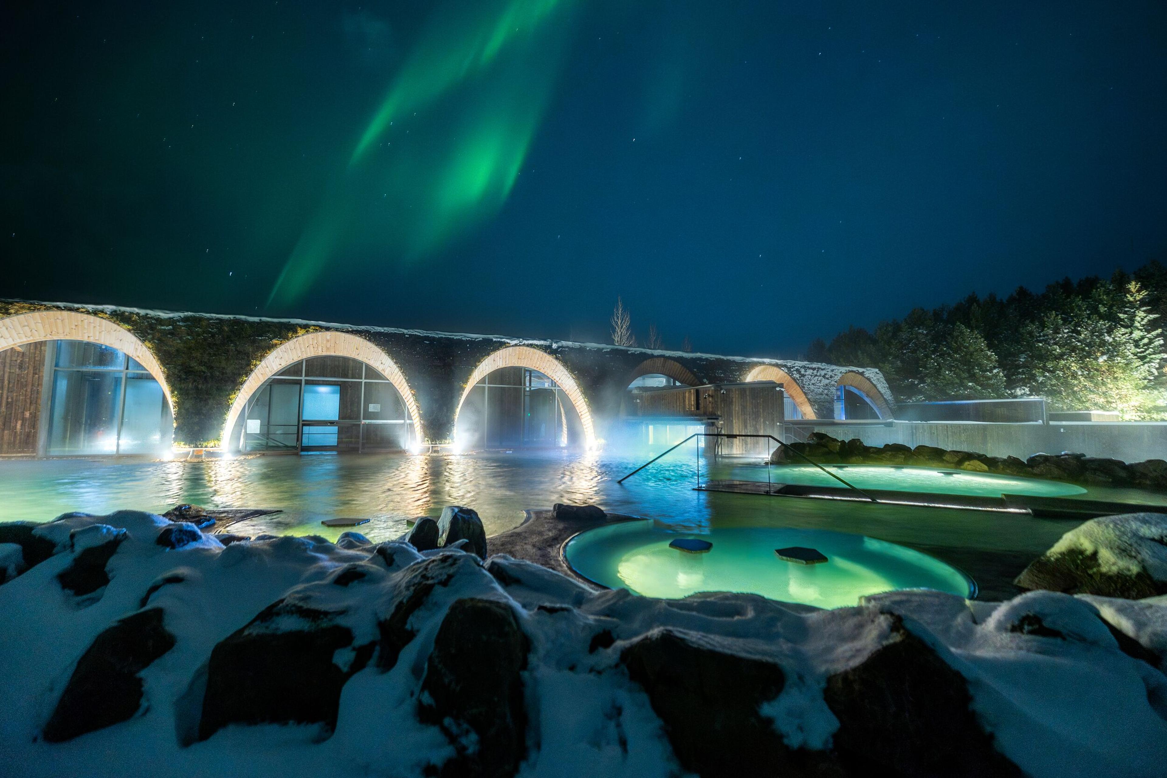Green Northern Lights above an arched building and steaming geothermal pools in a snowy night scene.