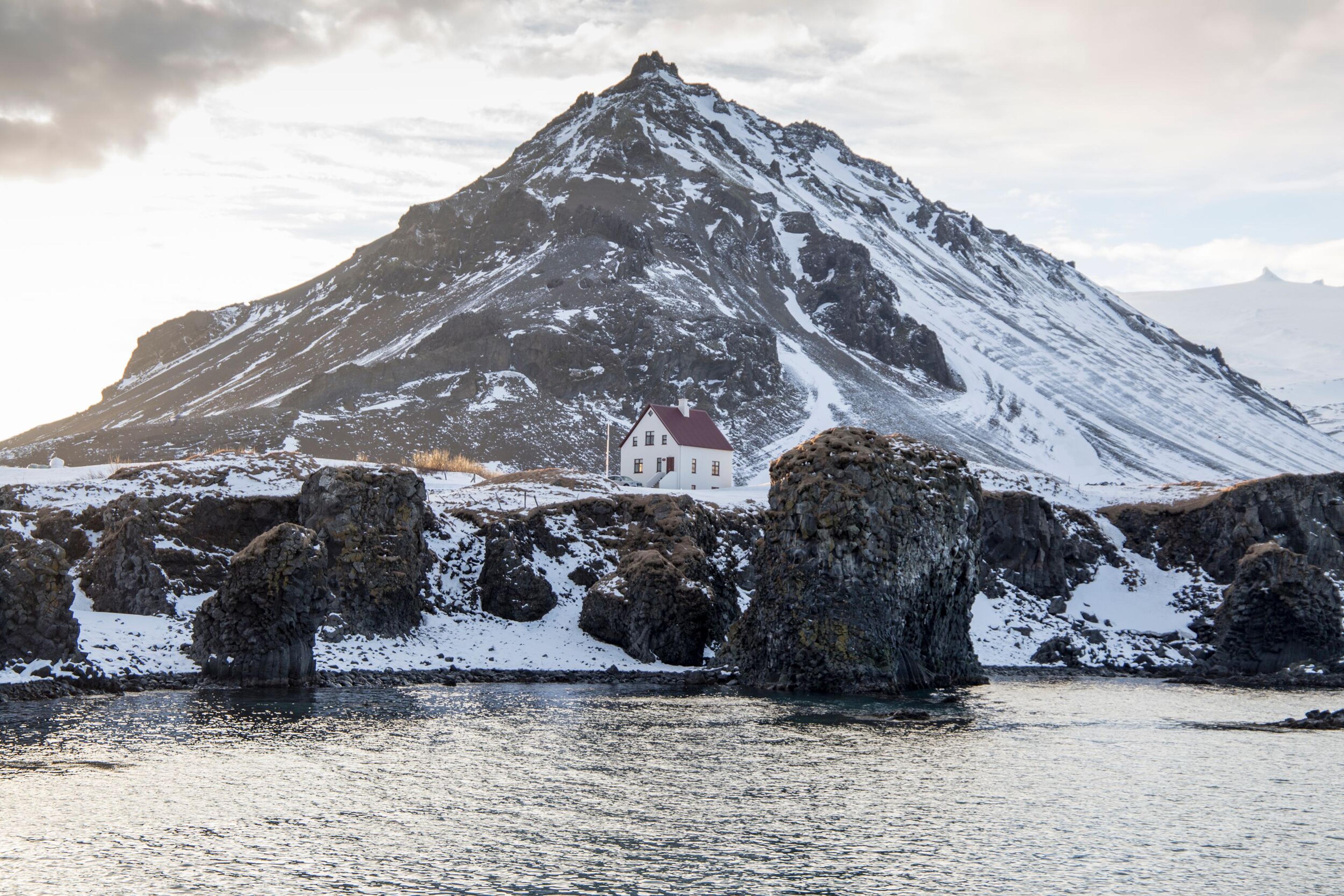 A white house with a red roof sits on a snowy, rocky coast with sea stacks, backed by a snow-covered mountain.