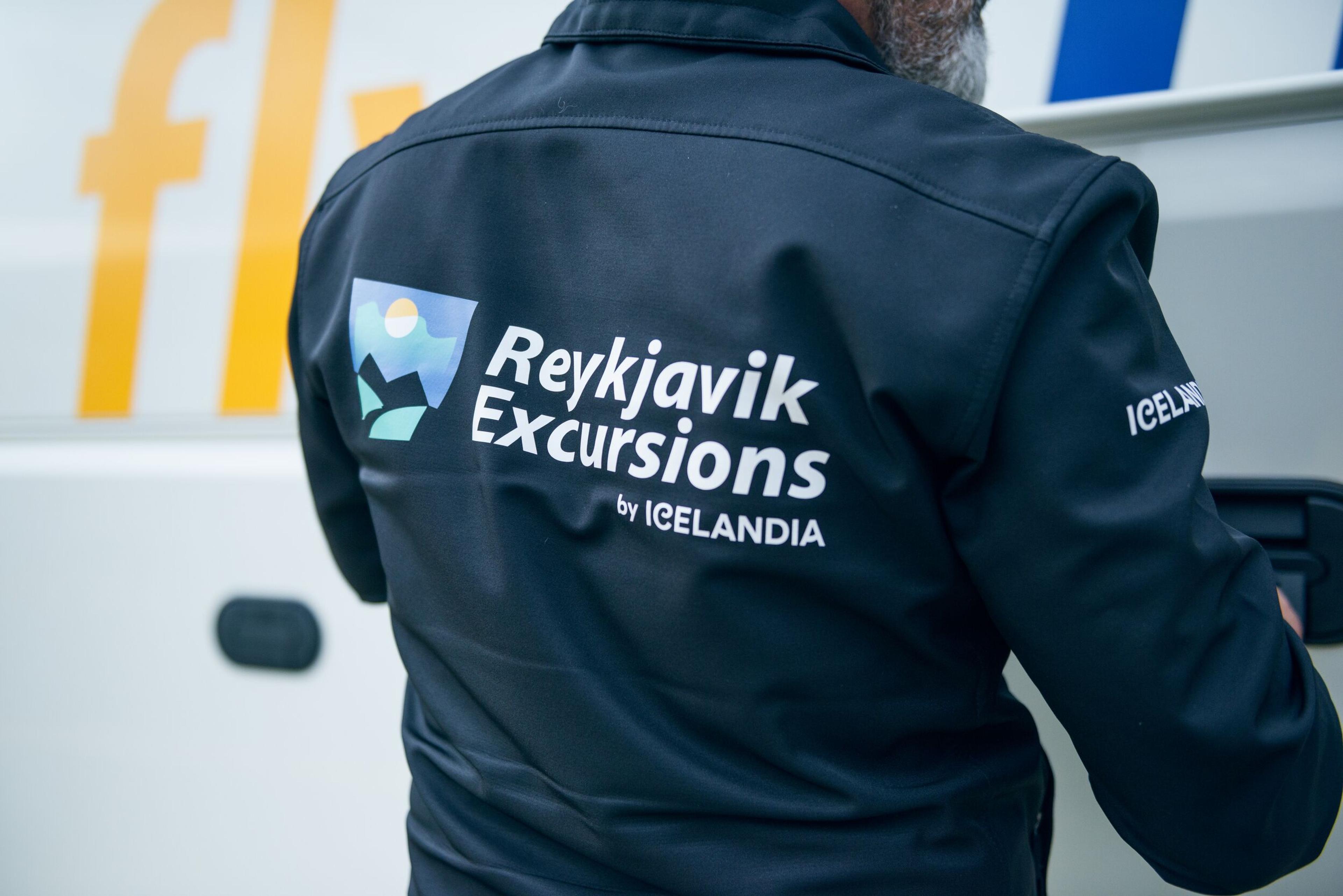 : Person wearing a "Reykjavik Excursions by Icelandia" jacket, standing near a tour bus, highlighting the excursion branding.