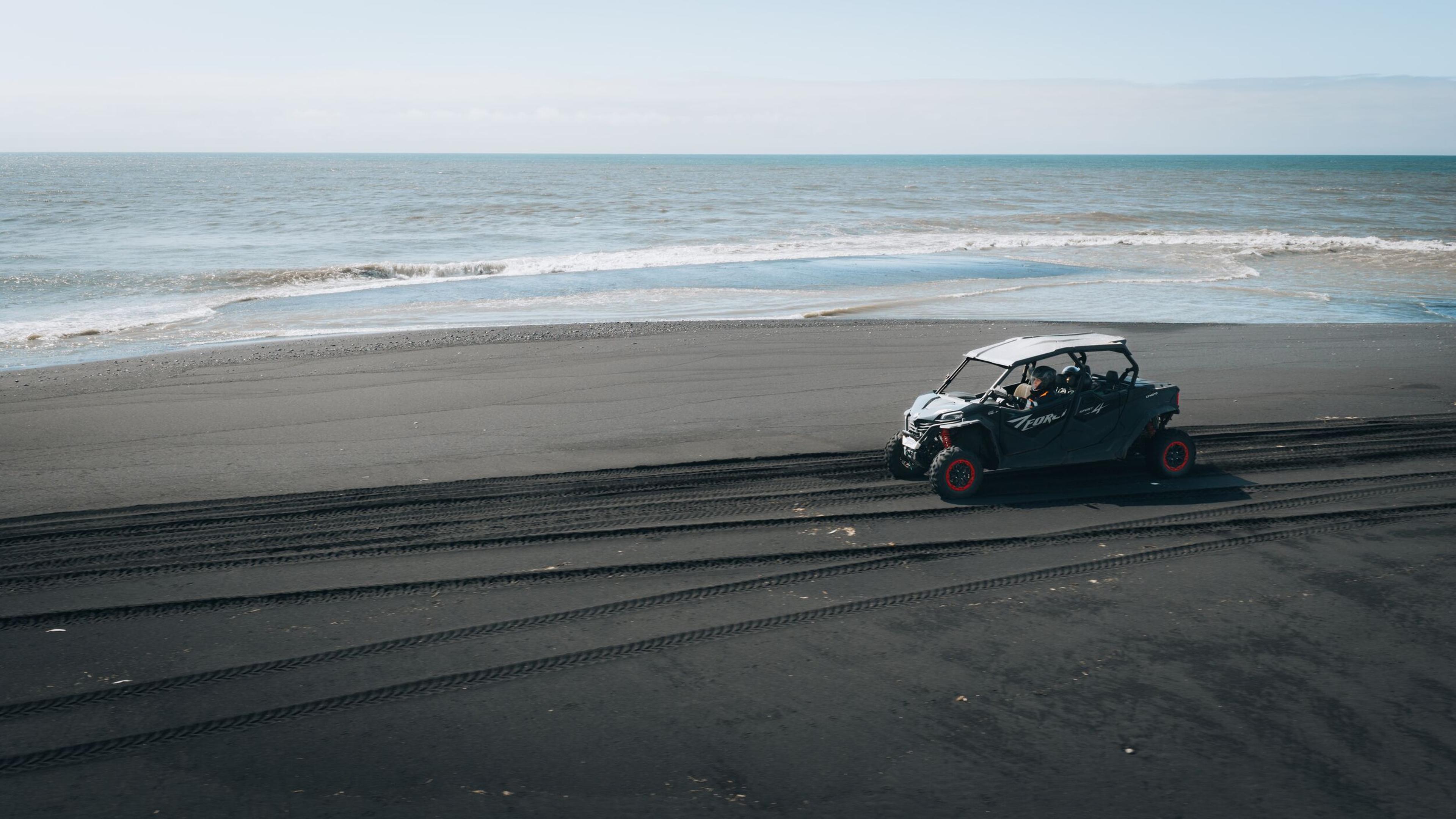 A gray ATV with red wheels drives on a black sand beach, leaving tracks, with the ocean in the background.