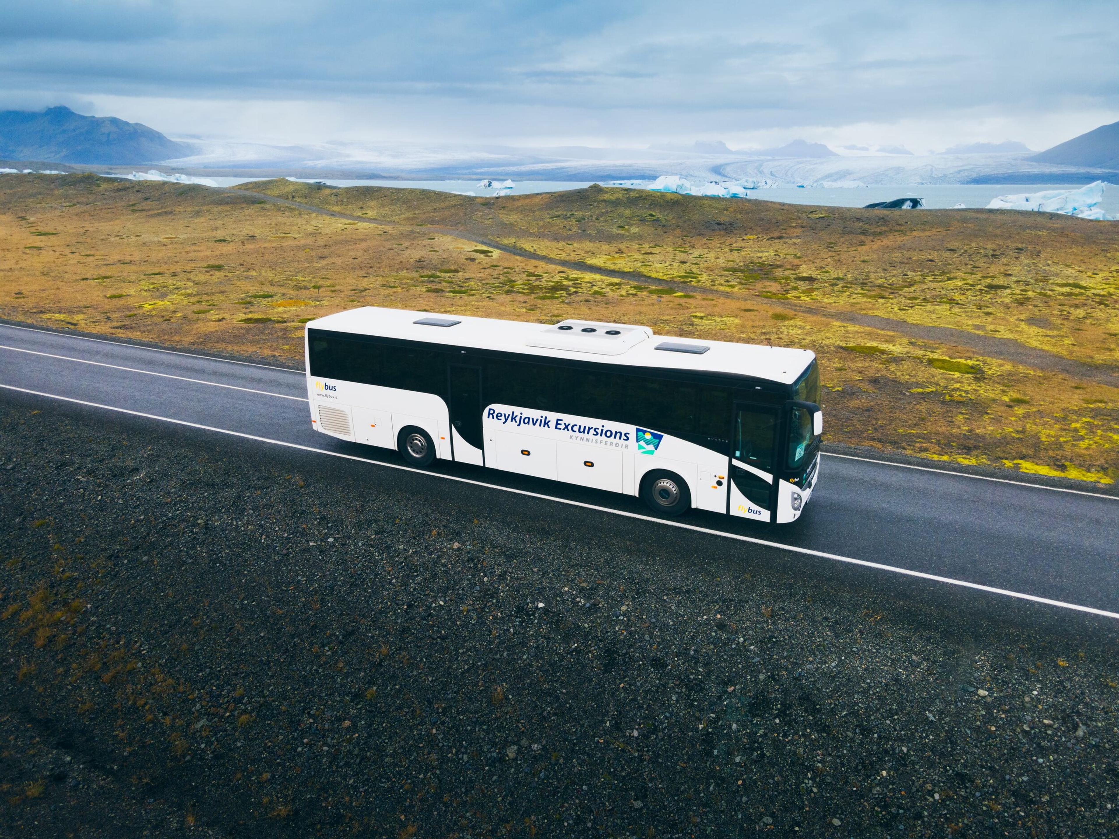 A tour bus traveling on a road through a vast, open landscape with mountains in the distance and overcast skies above.