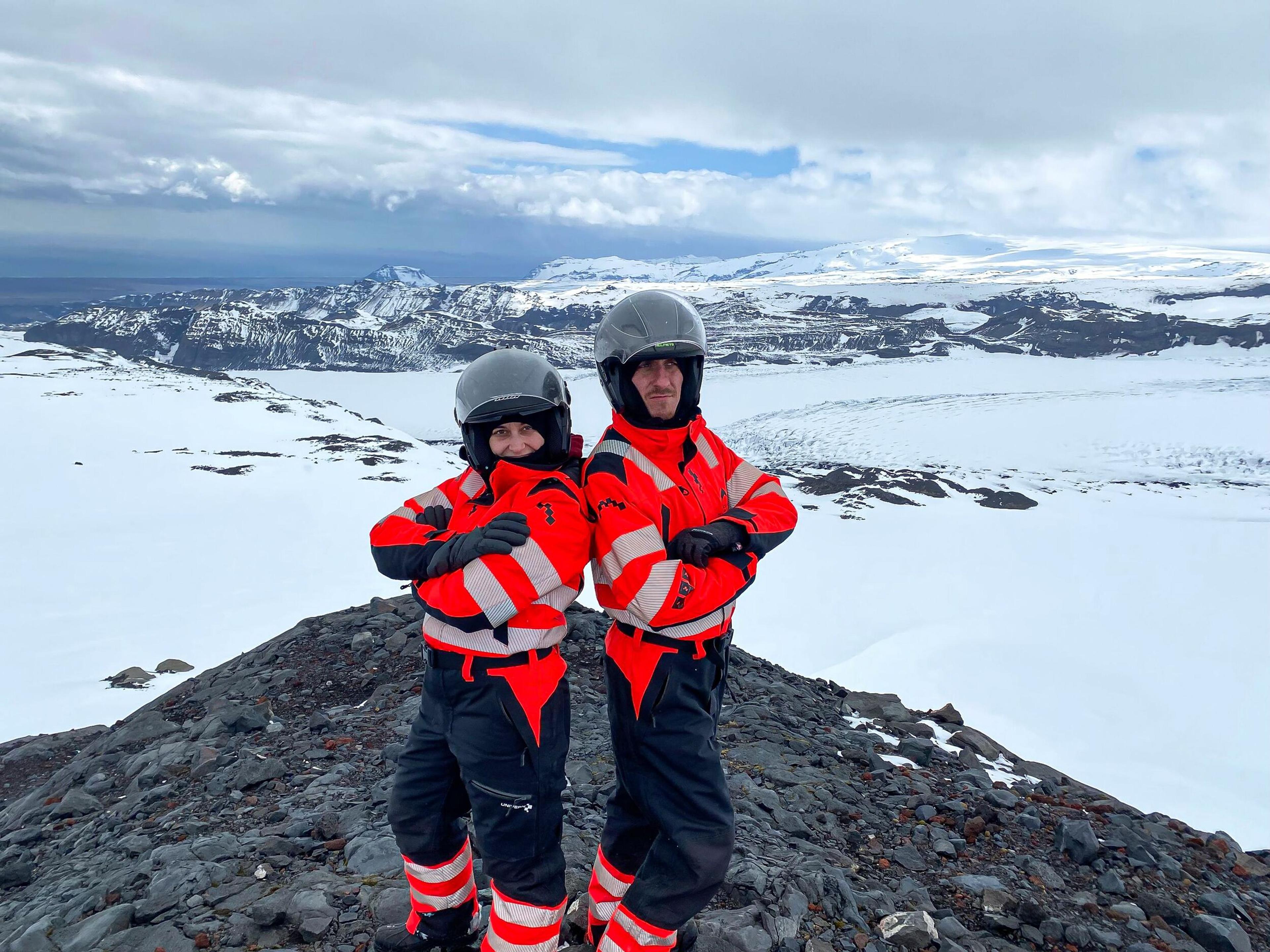 Two people in orange safety suits and helmets taking standing on a snowy overlook with mountains in the distance.