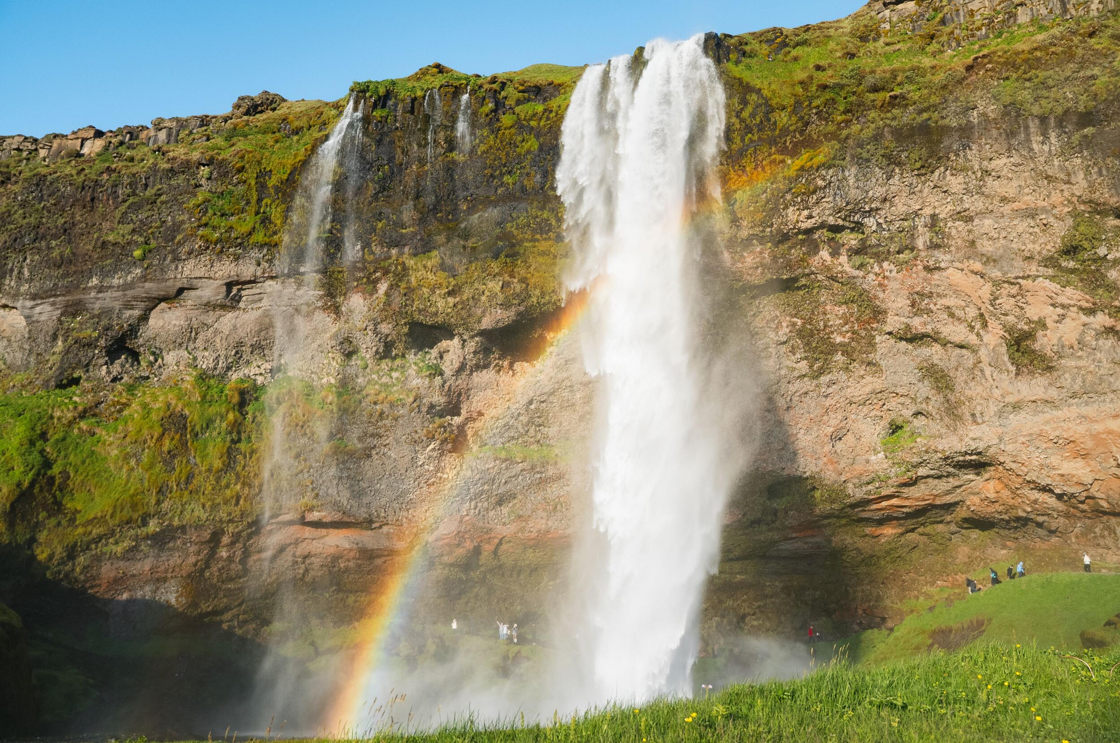 A powerful waterfall plunges from a green, rocky cliff, with a rainbow forming in its mist. People stand near the base.