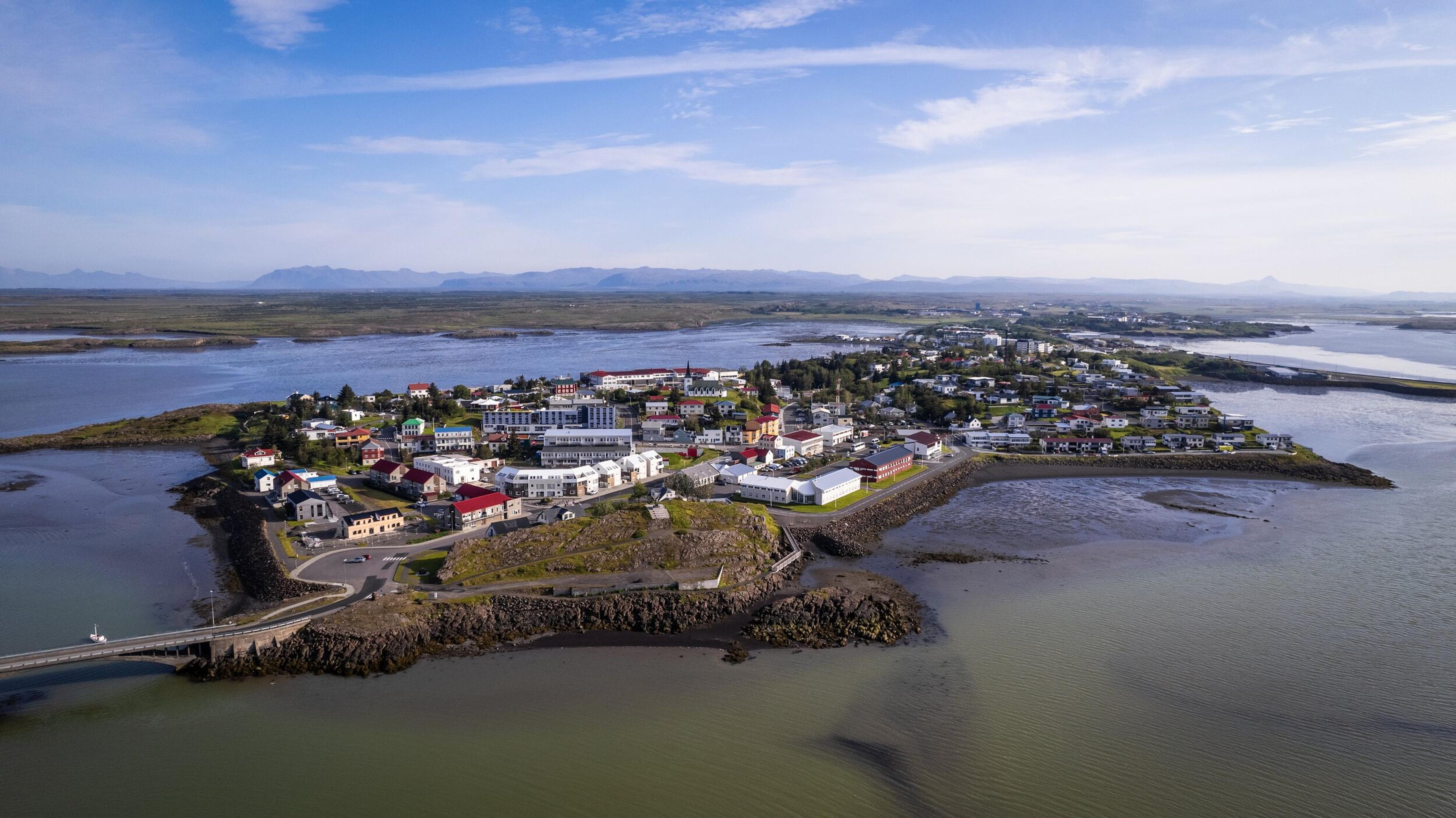 Aerial view of Borgarnes, a picturesque coastal town in Iceland surrounded by calm waters, with colorful buildings and a backdrop of distant mountains under a clear blue sky.