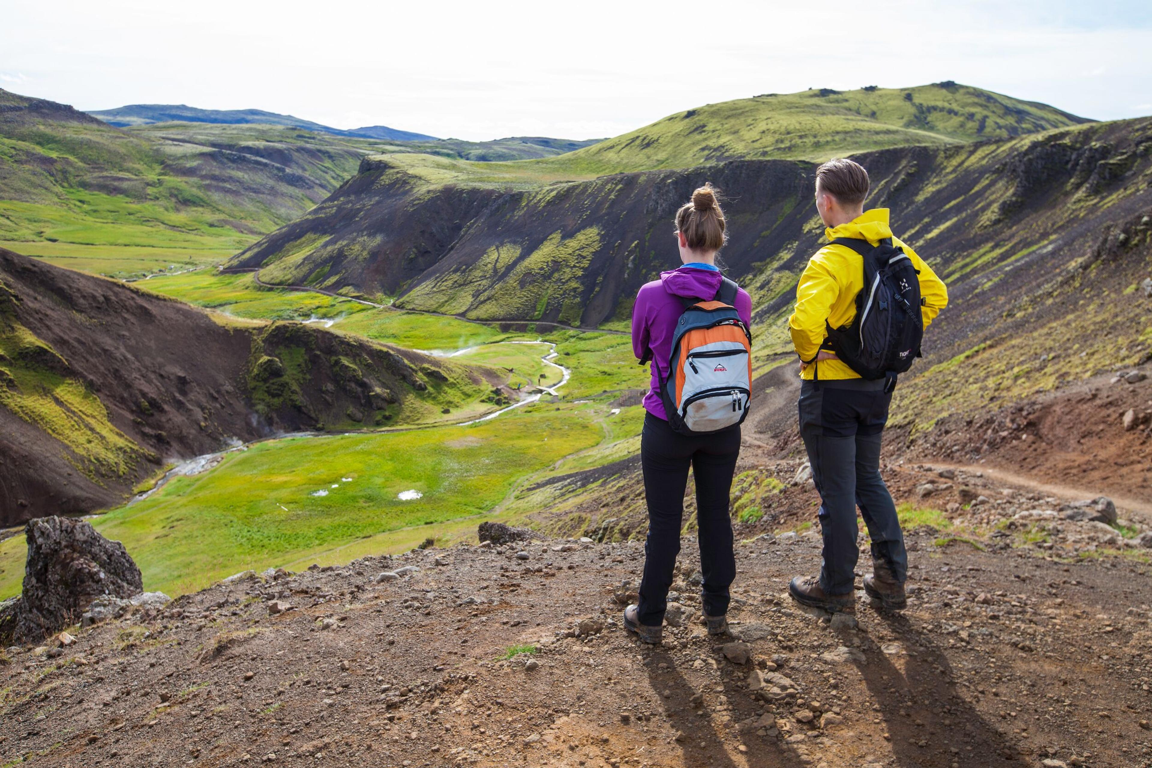 Two hikers with backpacks overlook a green valley with a winding river.