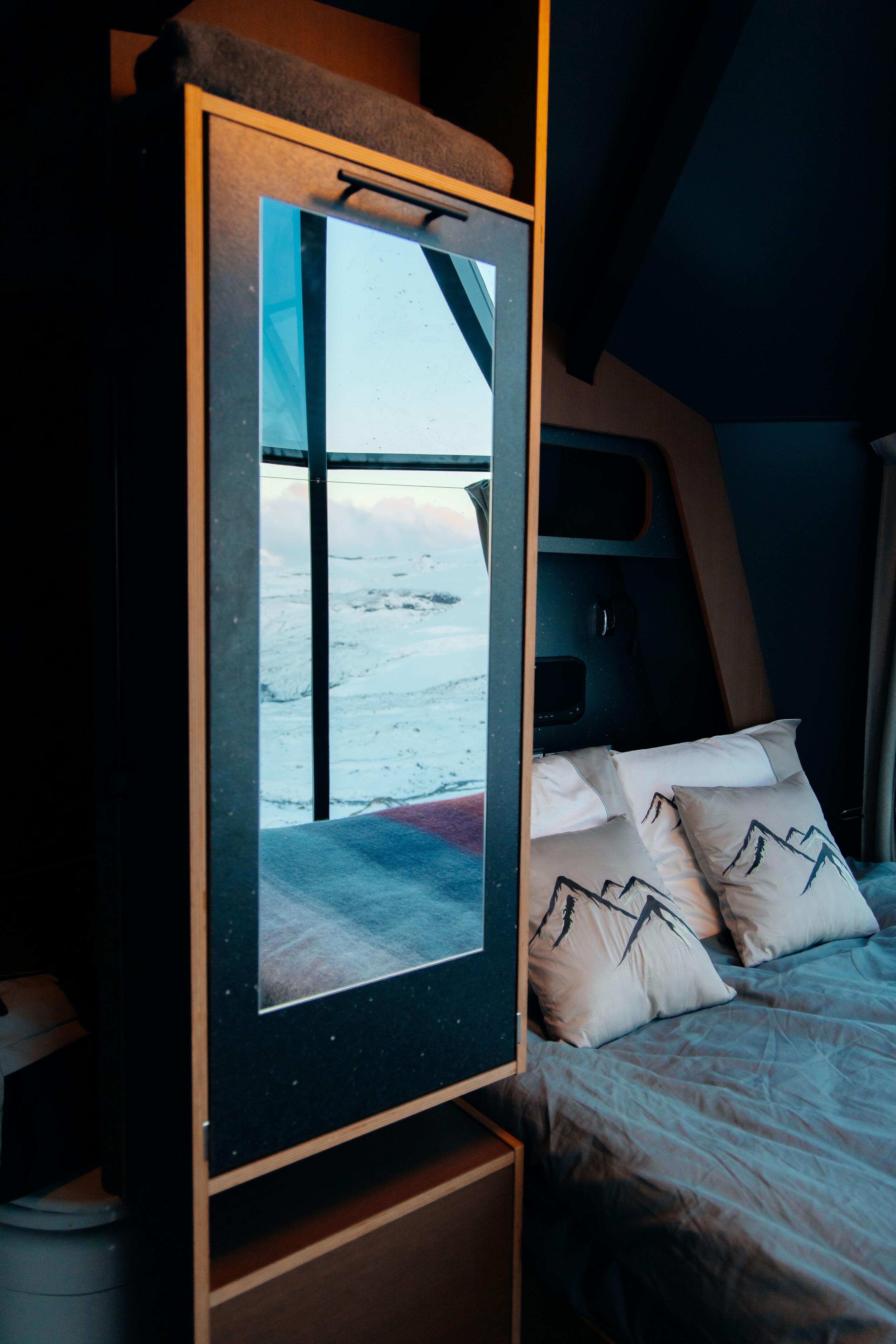 A bedroom interior with a bed, mountain-print pillows, and a tall mirror reflecting a snowy landscape.