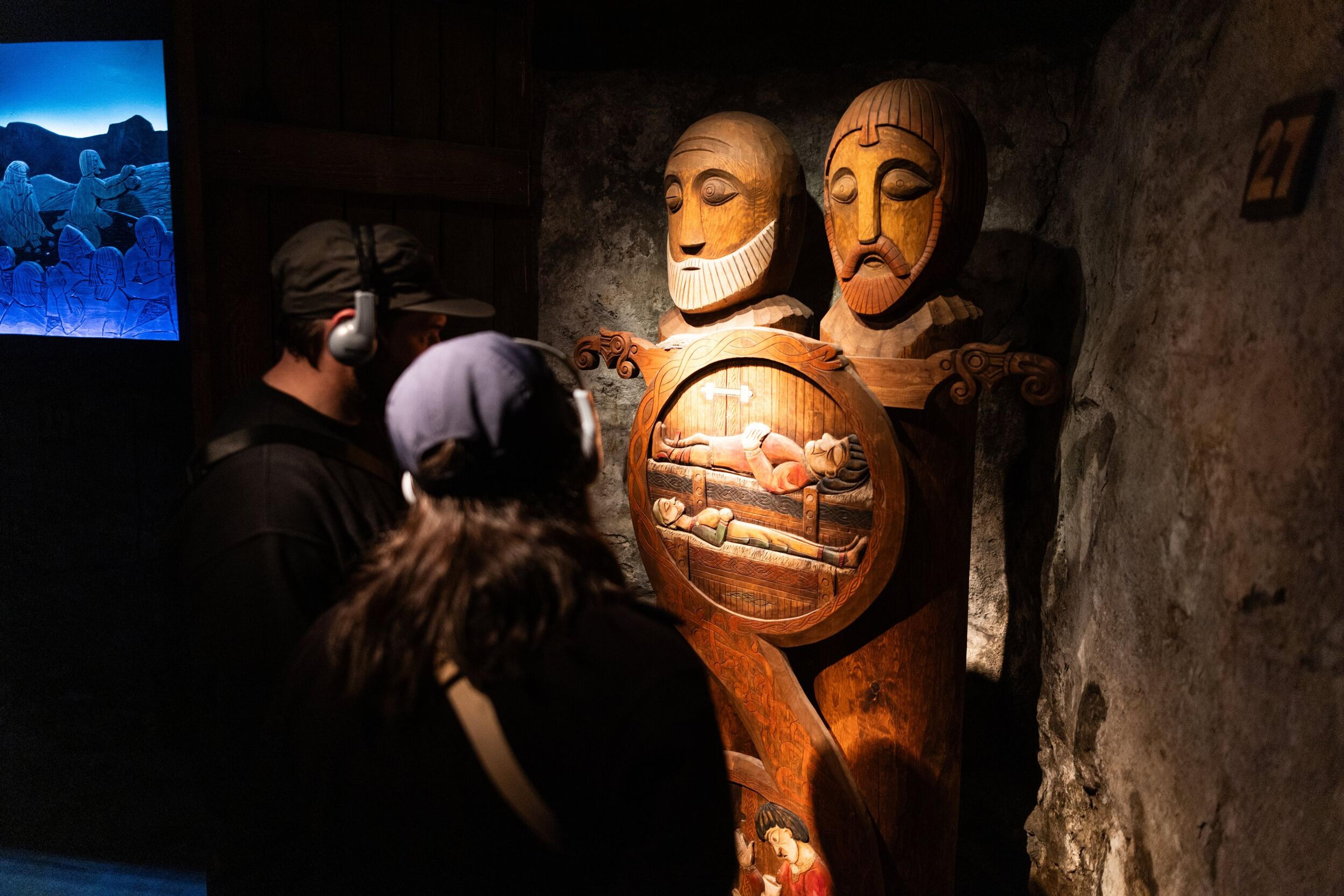 Visitors observing a wooden sculpture depicting two historical figures with intricate carvings at the Settlement Exhibition in Borgarnes, showcasing Iceland's cultural heritage in a dimly lit setting.