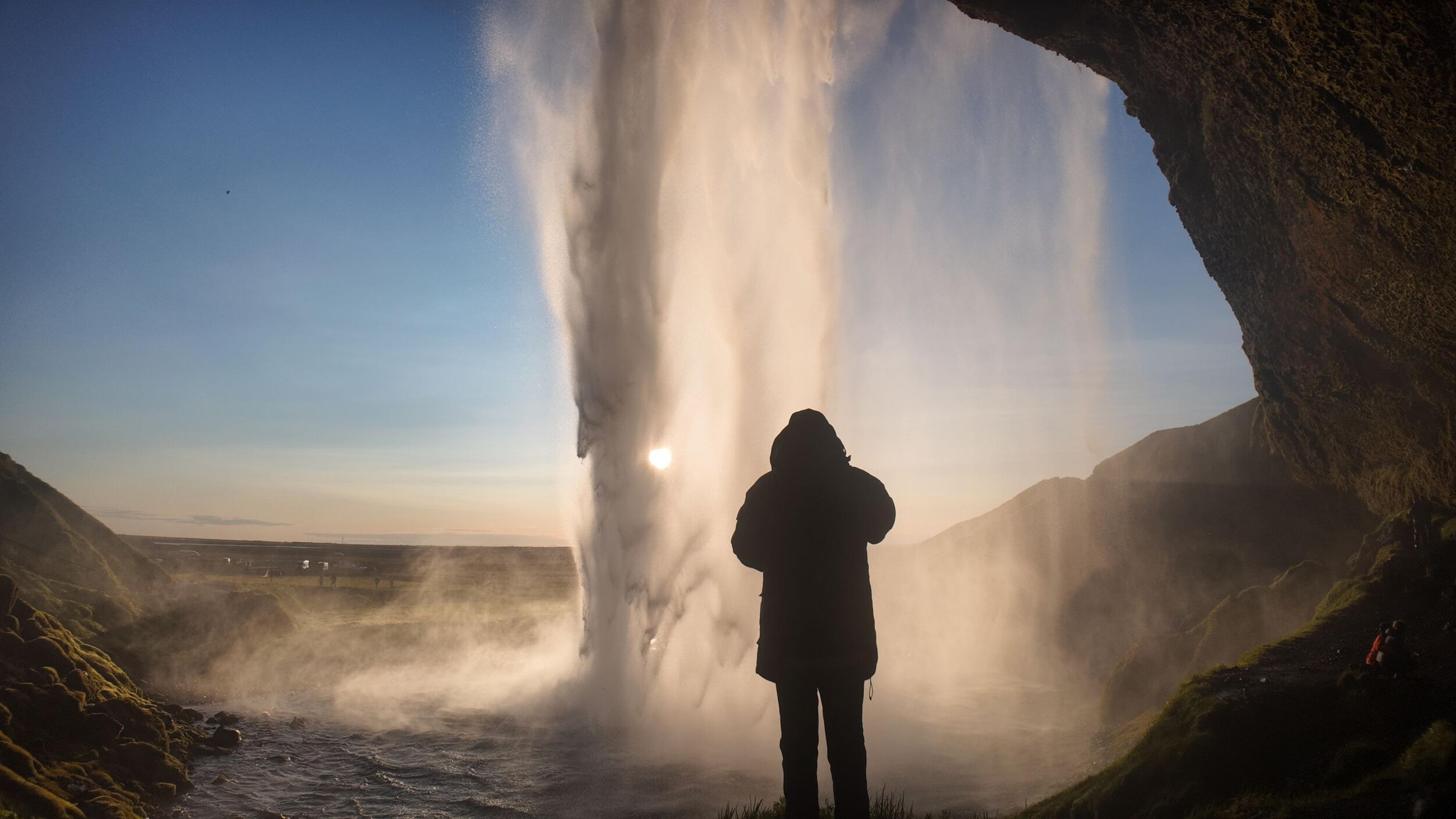 Silhouetted person standing behind a powerful waterfall backlit by the sun.