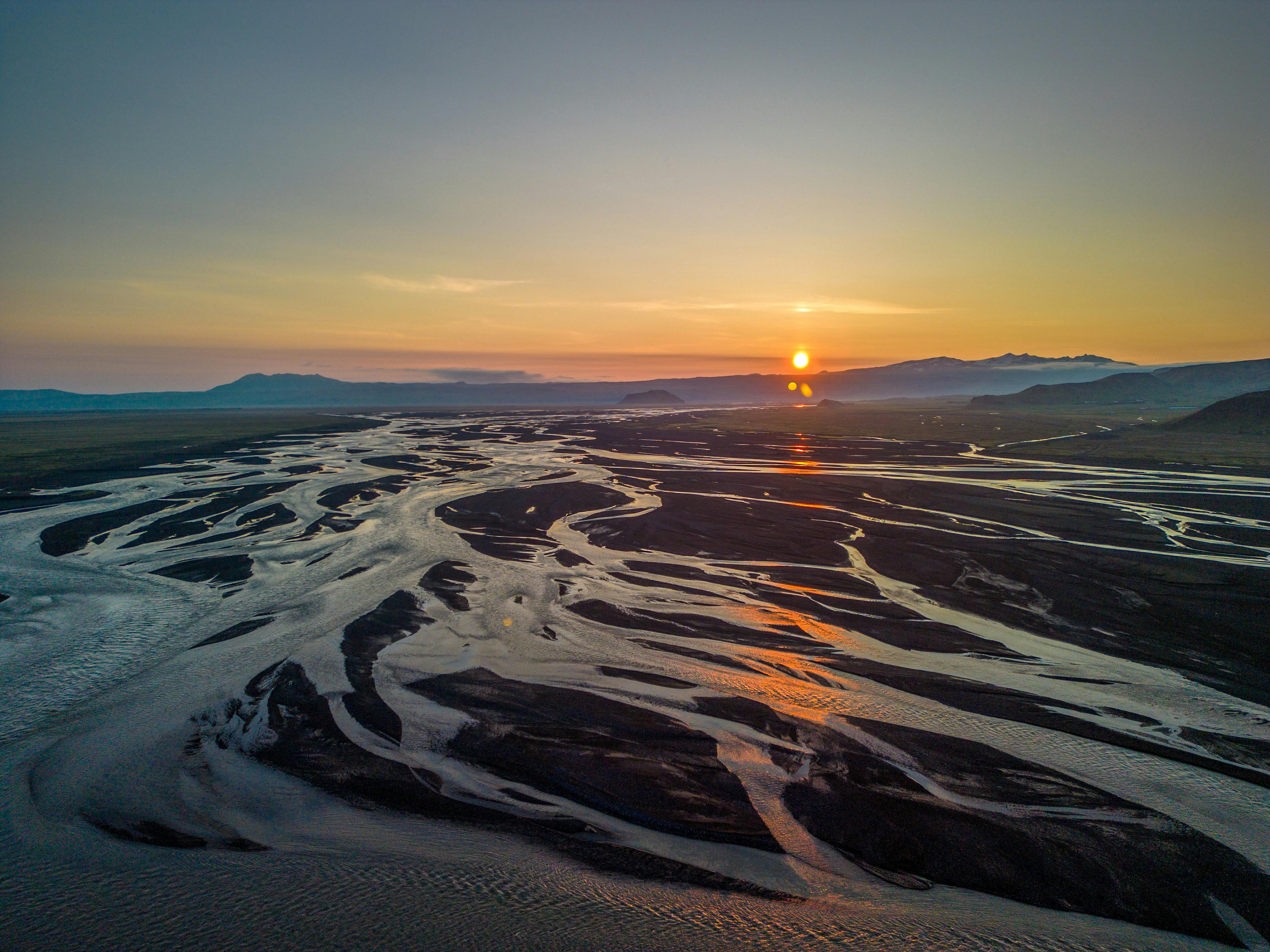 Aerial view of a braided river flowing through a vast landscape at sunset, with the sun dipping behind distant mountains.