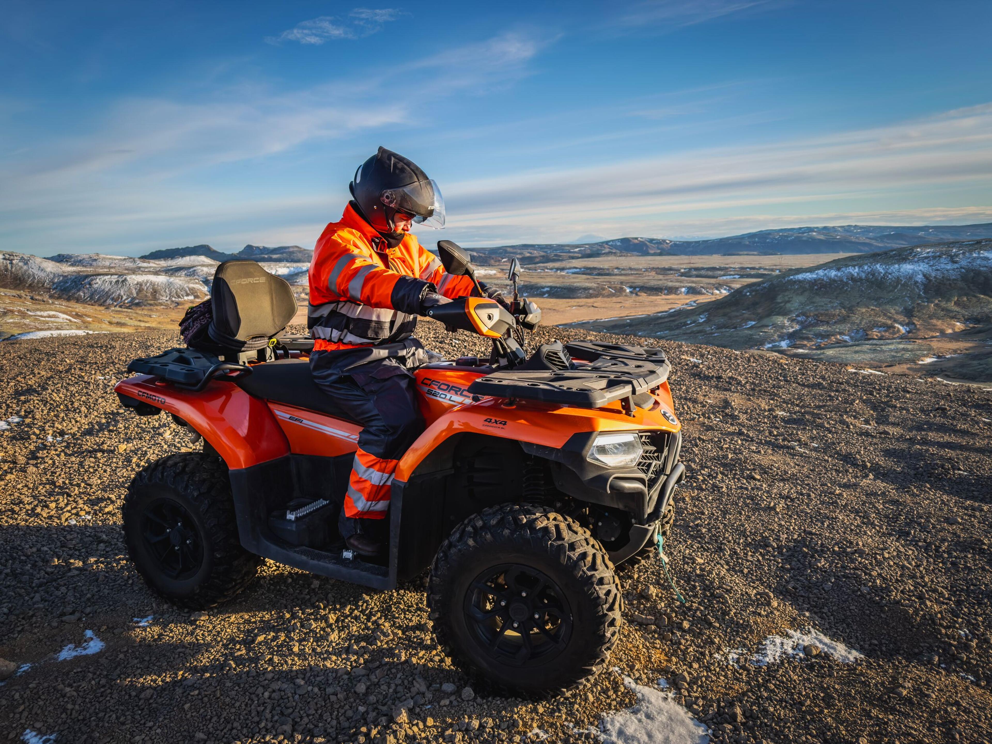 Person in a helmet and orange reflective suit on an orange ATV on a rocky hill overlooking a snowy landscape.