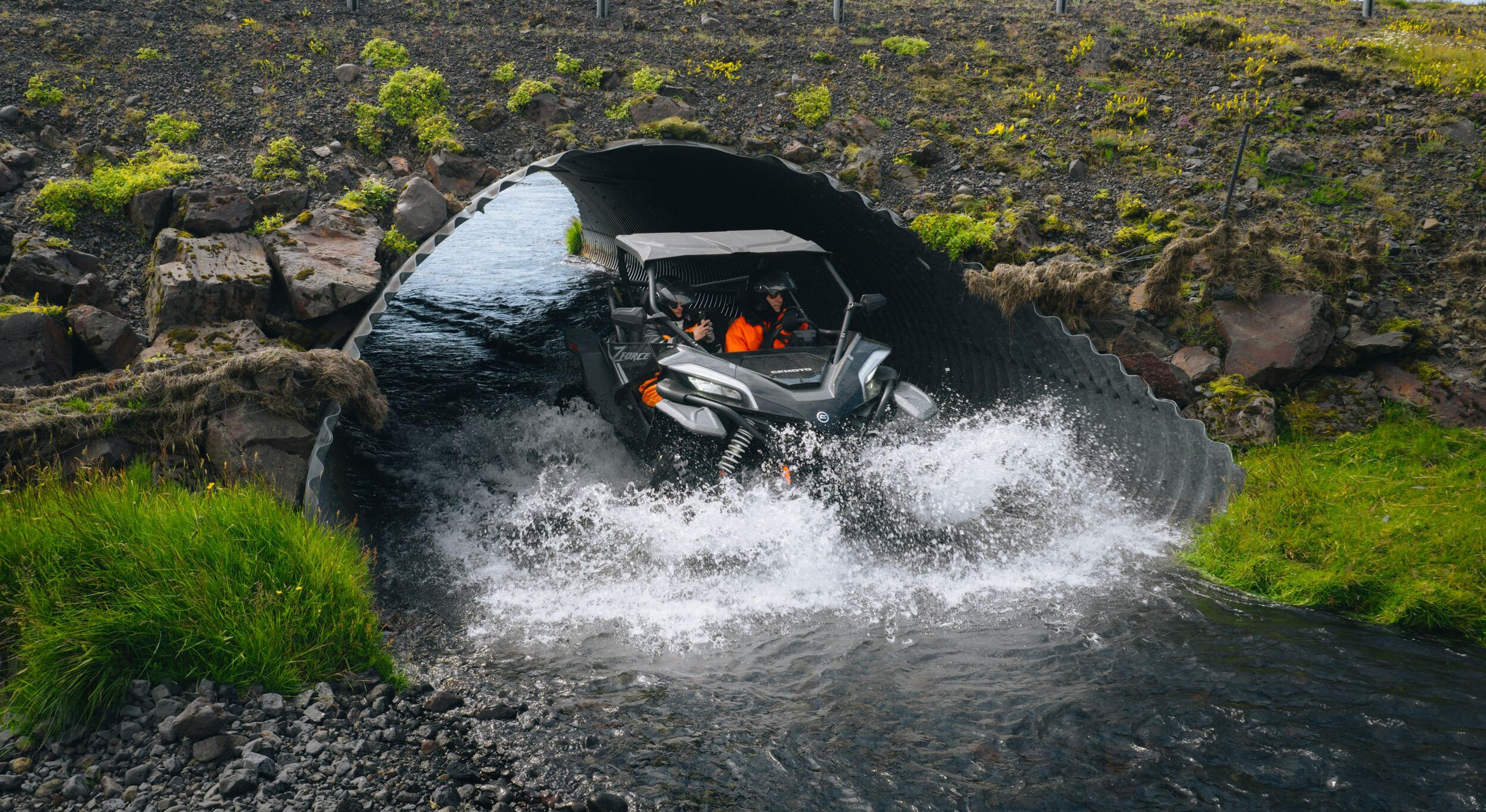 A side-by-side vehicle with two occupants drives through a stream, splashing water as it exits a metal culvert.