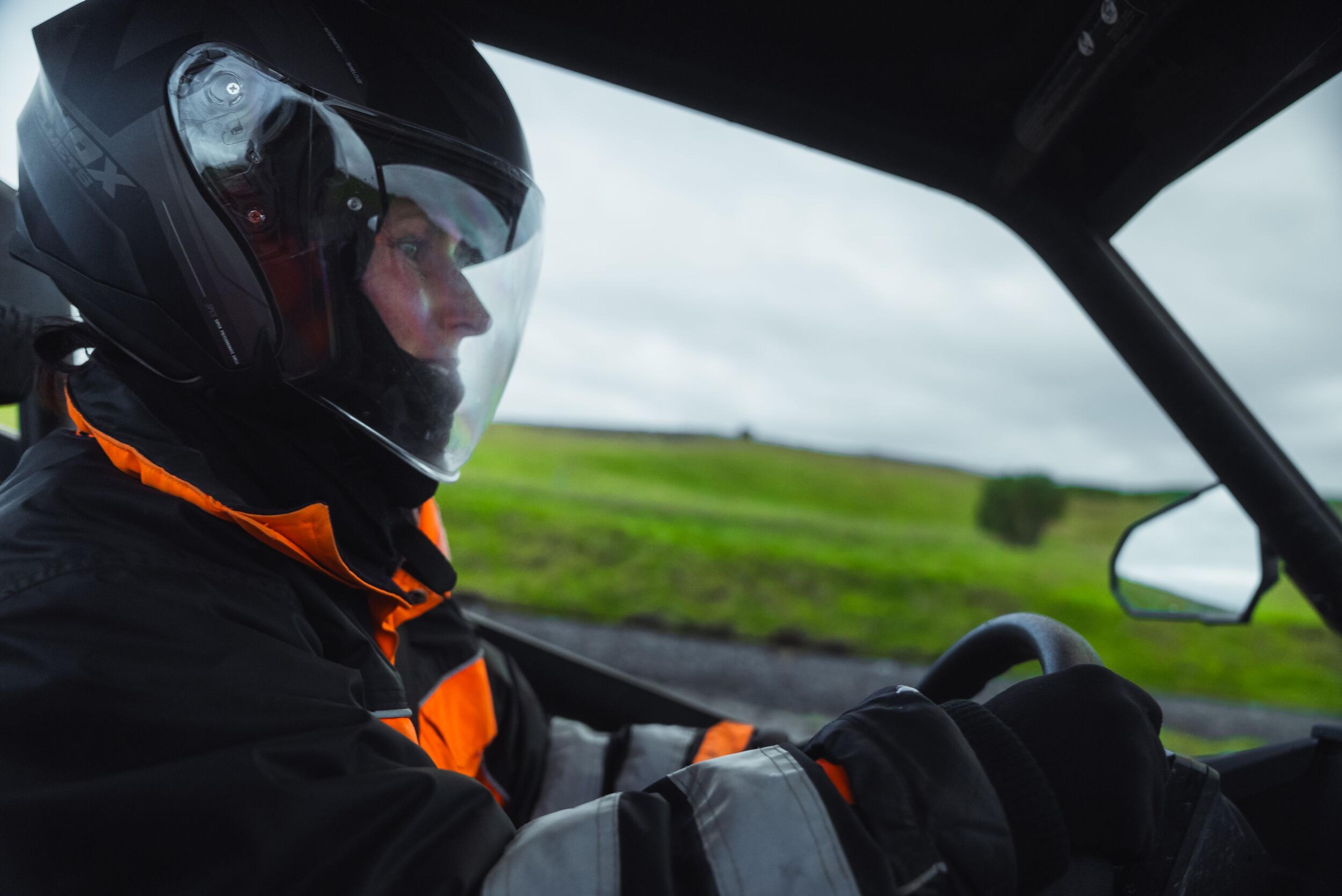 Driver in a black helmet and orange jacket steering an off-road vehicle with green hills in the background.