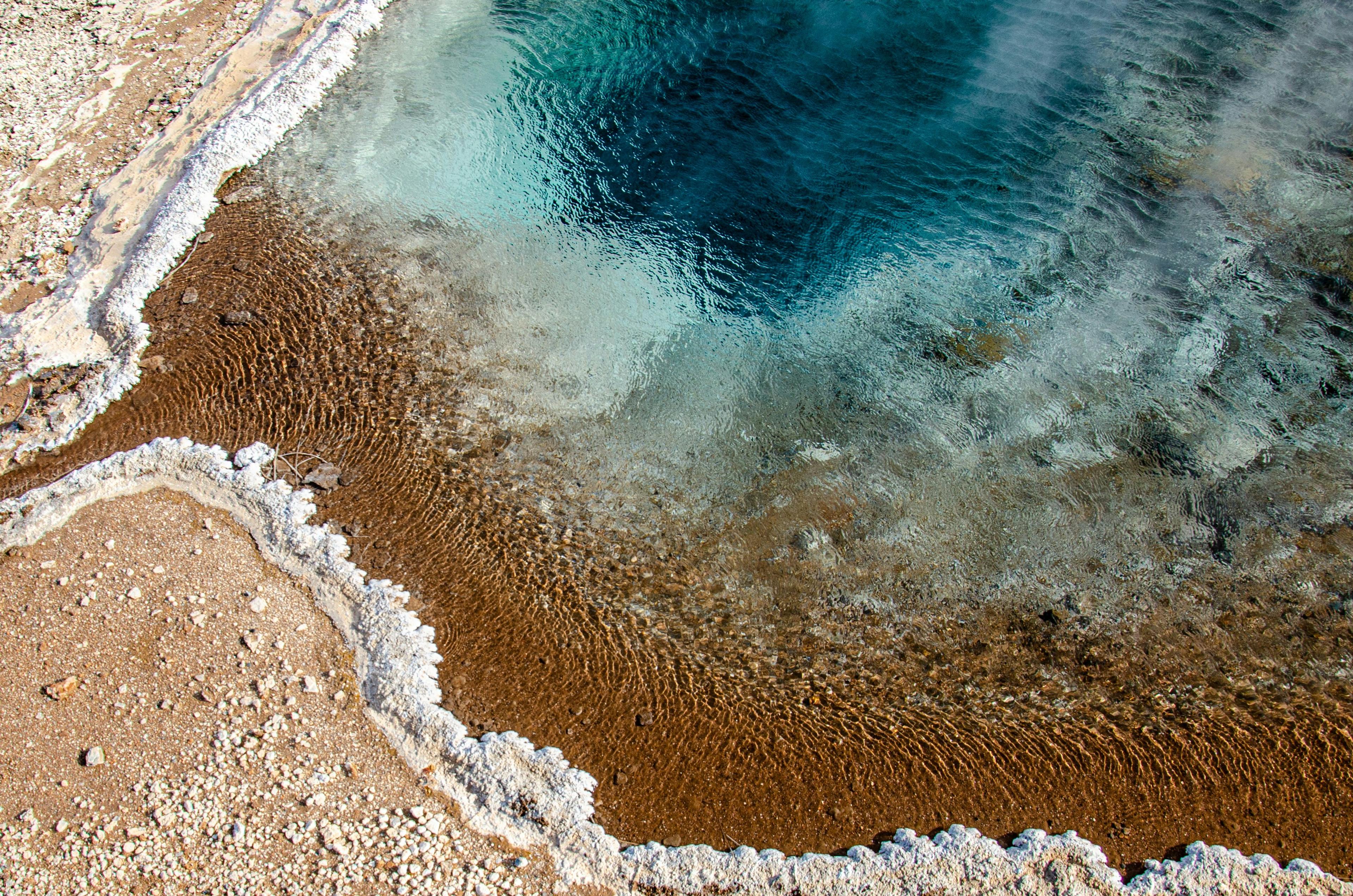 The photo depicts a close-up of a geothermal hot spring, showcasing the mesmerizing transition from the clear blue water at its center to the mineral-rich earthy tones and crystalline textures at its edges.