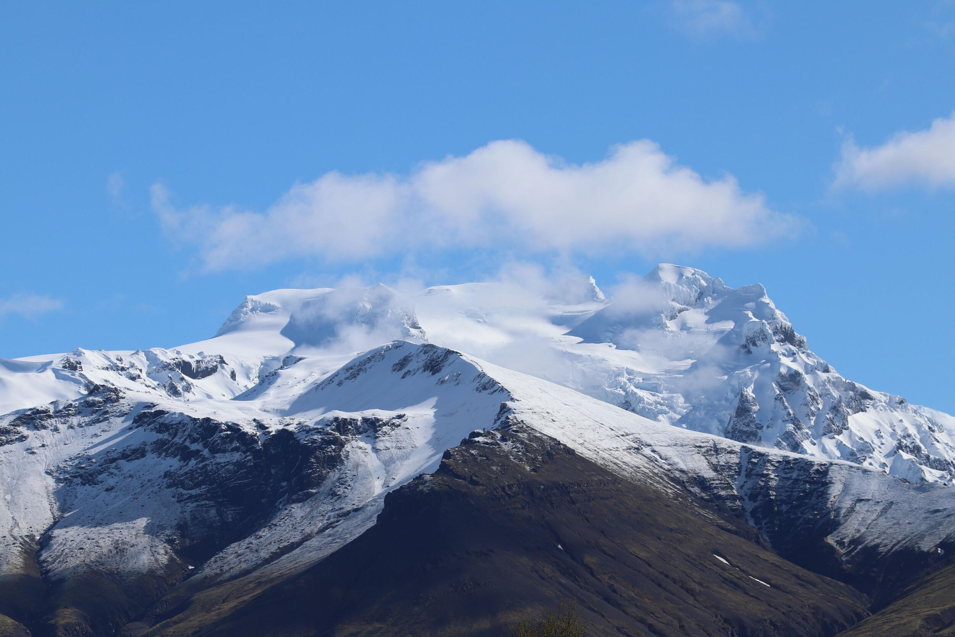 A snow-covered mountain with glaciers and dark rocky lower slopes, under a blue sky with scattered clouds.