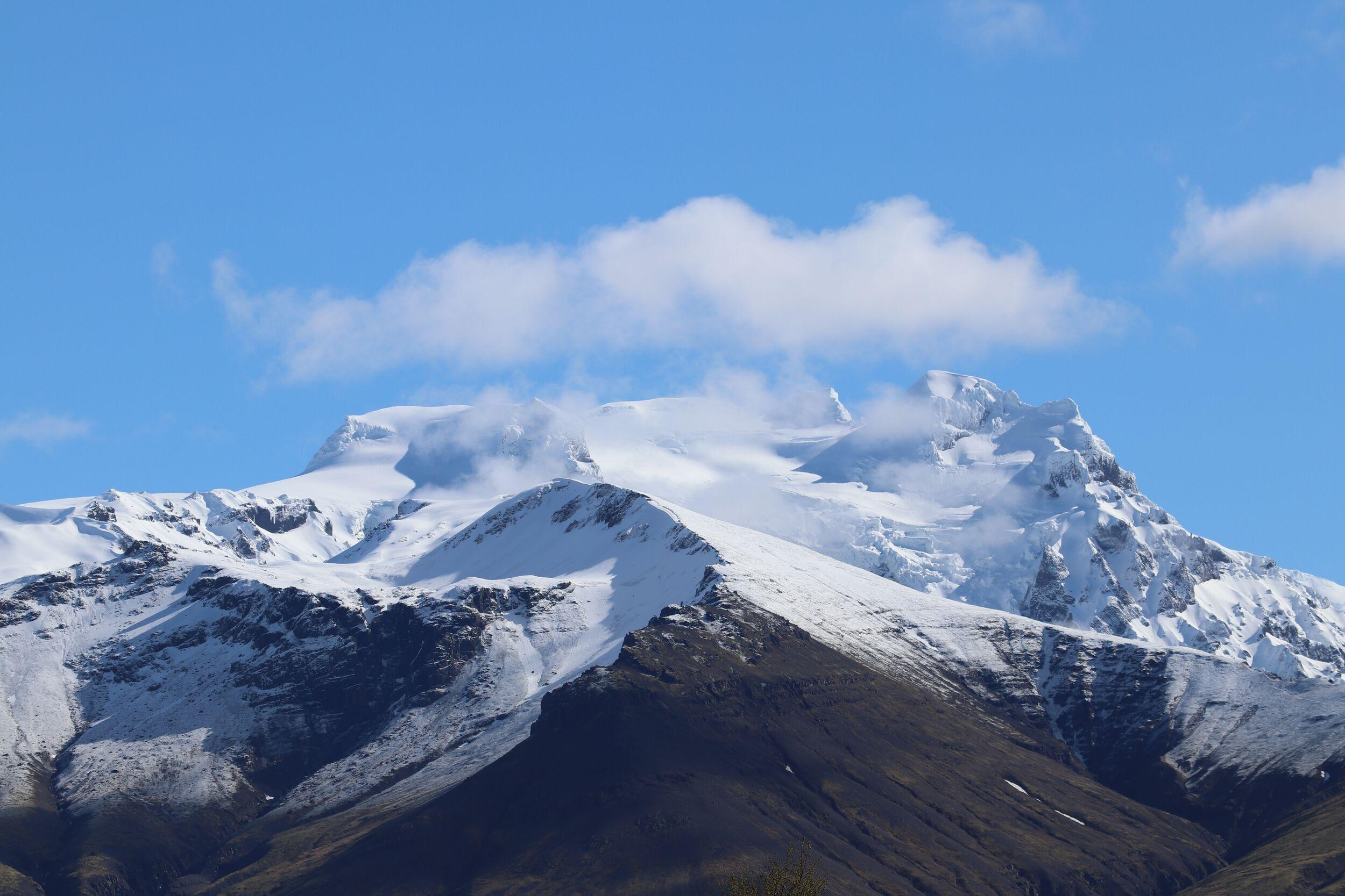 A snow-covered mountain with glaciers and dark rocky lower slopes, under a blue sky with scattered clouds.