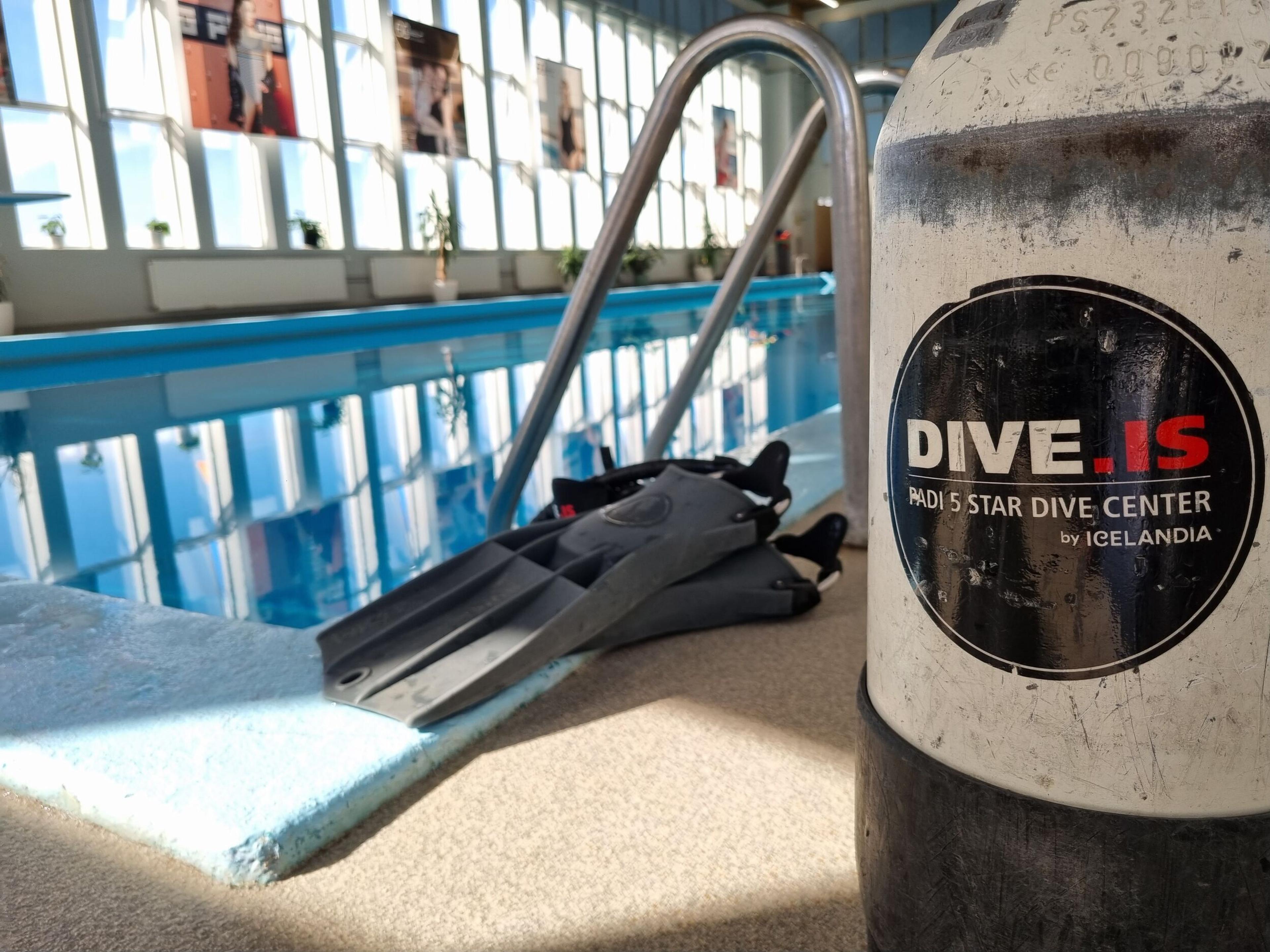 A DIVE.IS branded scuba tank and black flippers by an indoor swimming pool with large windows.