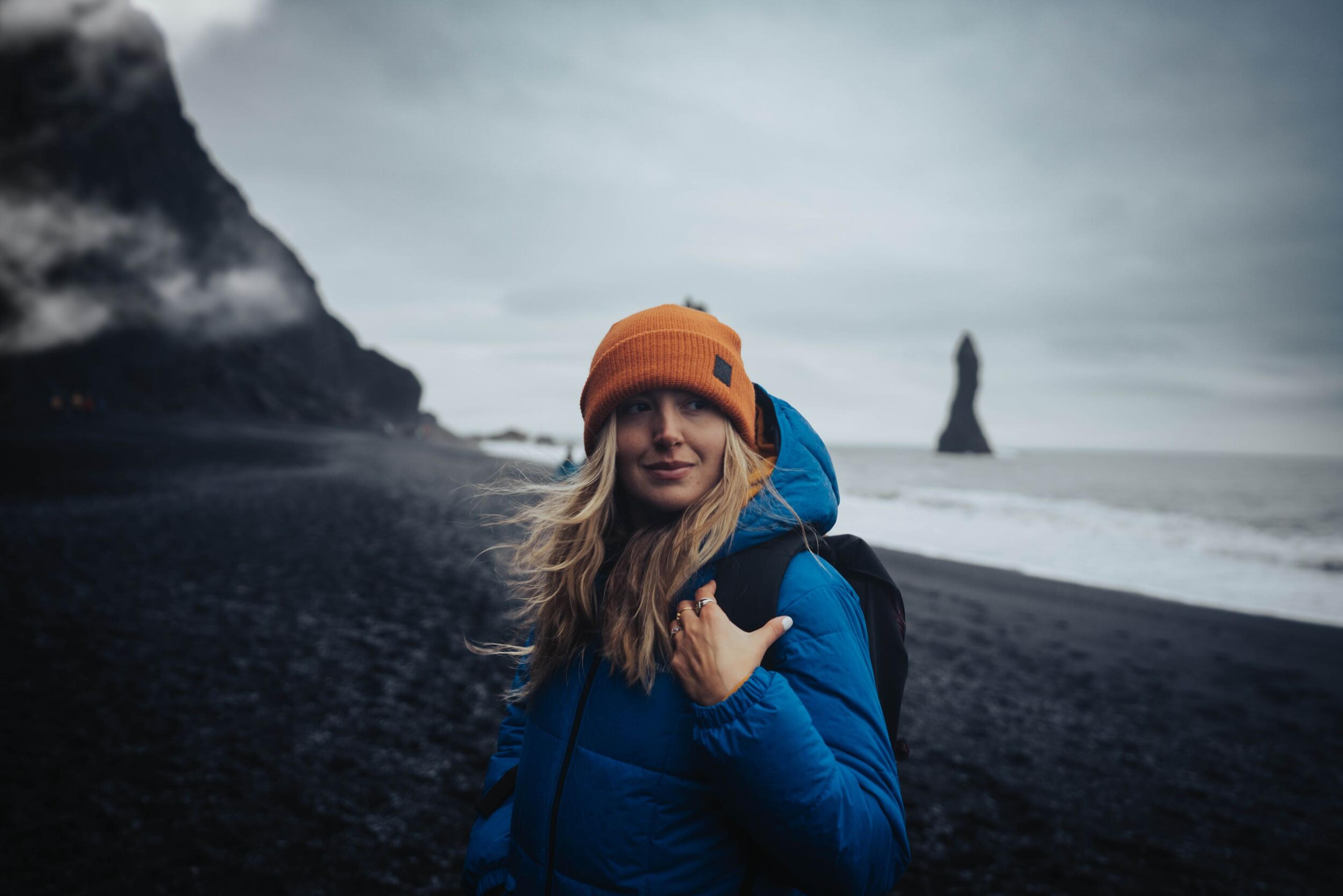 Blond, long-haired woman in orange cap and blue down jacket with Reynisfjara black sand beach in the background