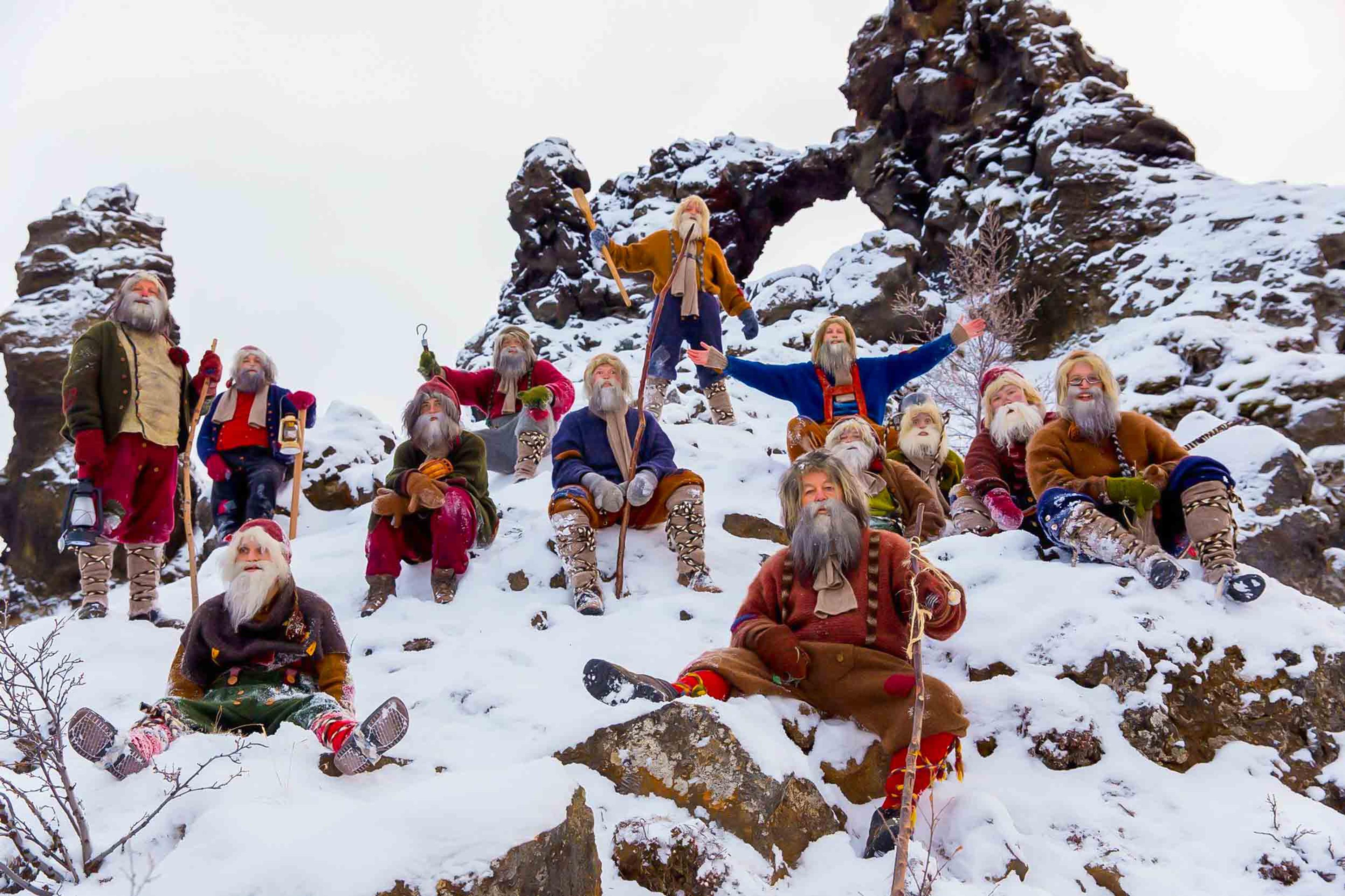 A group of whimsical figures resembling traditional Santa Clauses, each with unique colorful costumes and long beards, posed playfully on a snowy hill with rocky formations in the background, creating a festive and mythical tableau