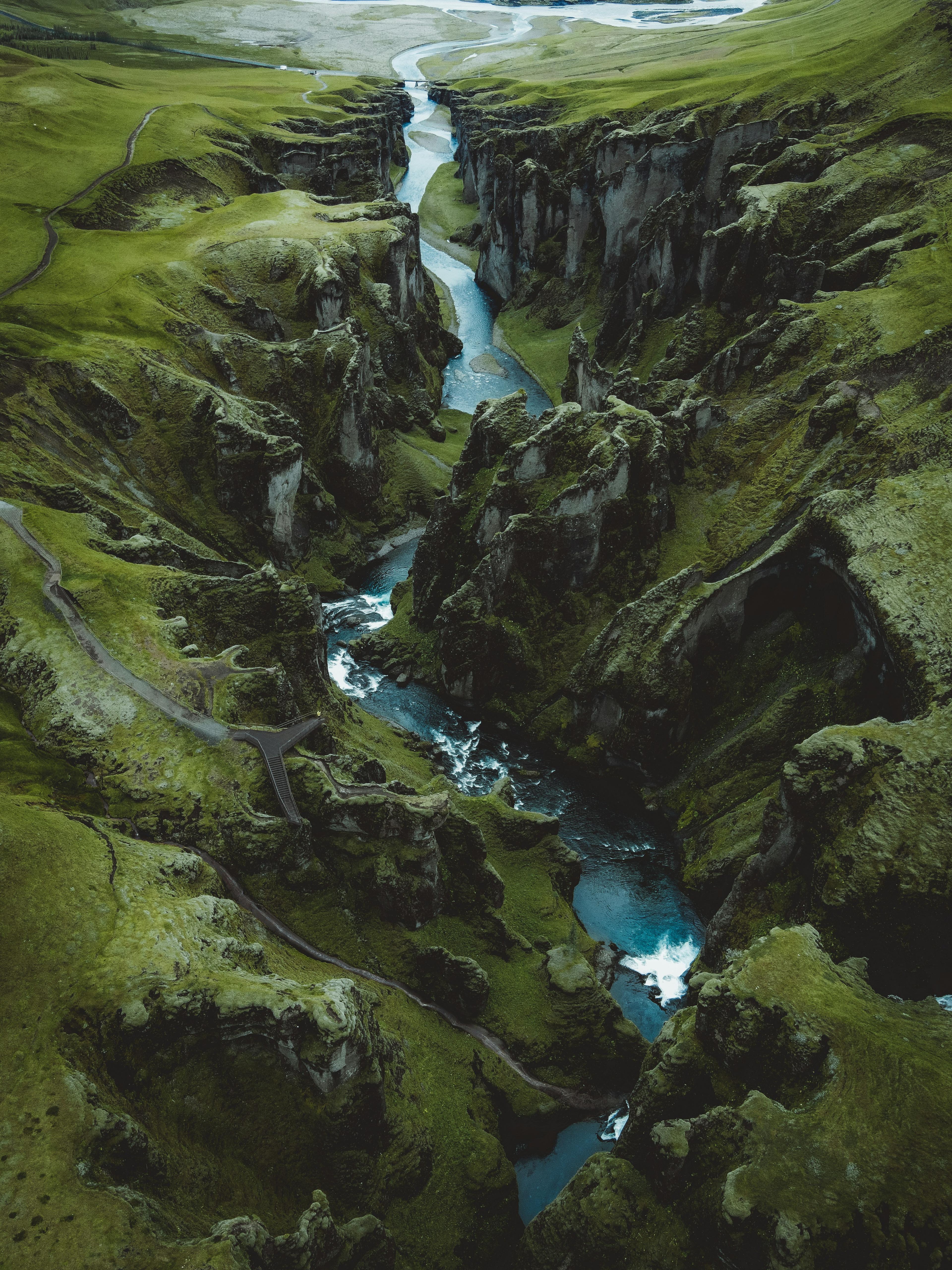 Aerial view of a dramatic, lush green canyon with winding turquoise rivers and waterfalls, carving through the rugged terrain under a cloudy sky.