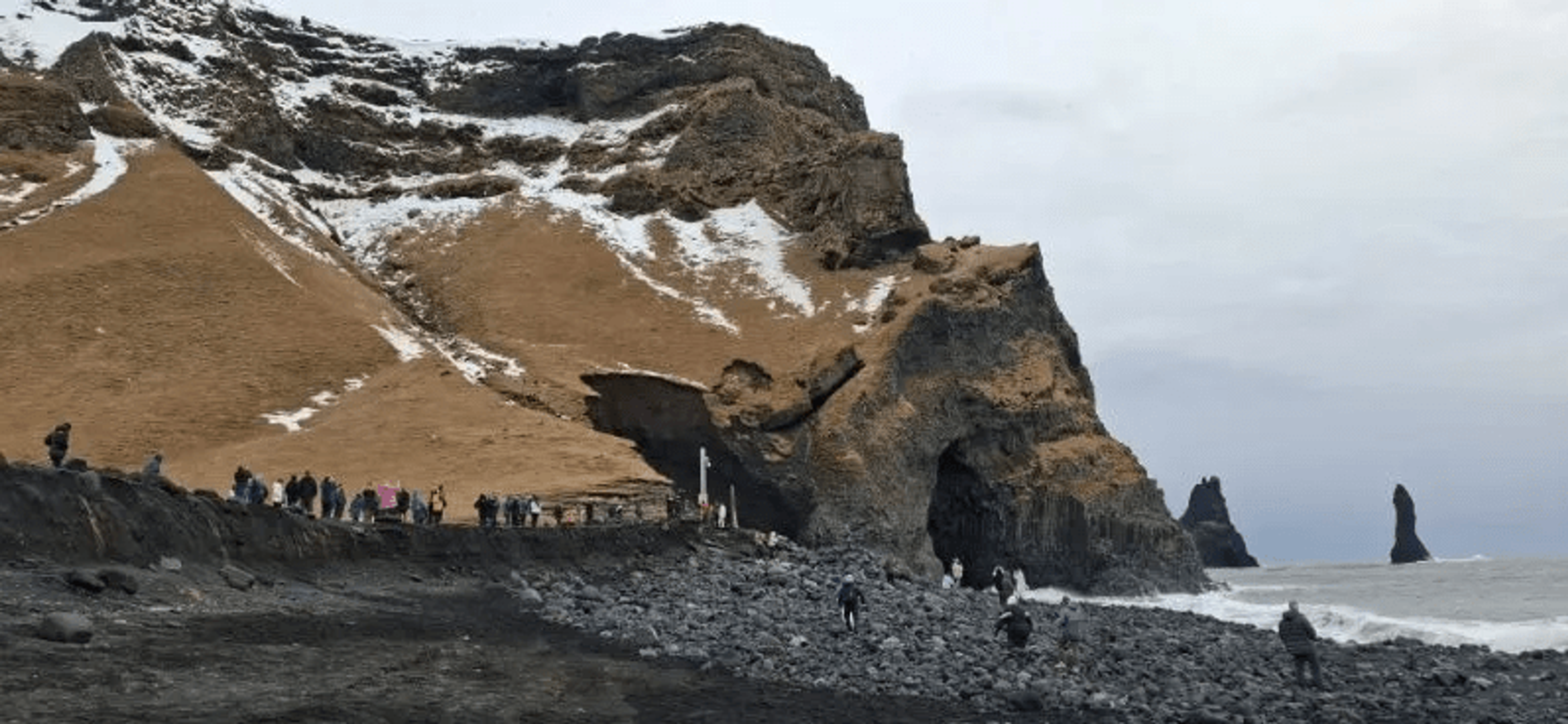 People on a black pebble beach below snow-dusted cliffs with a sea cave and ocean rock formations.
