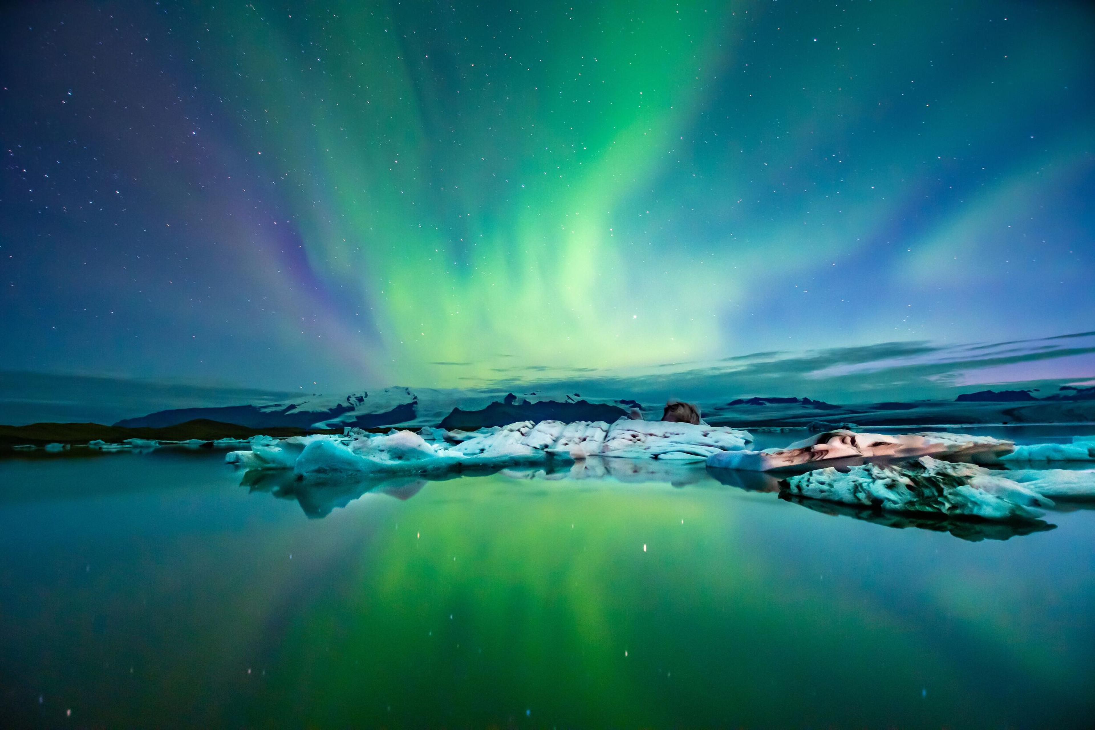 Green and purple aurora borealis over a lake with icebergs, reflecting on the water, with mountains in the background.