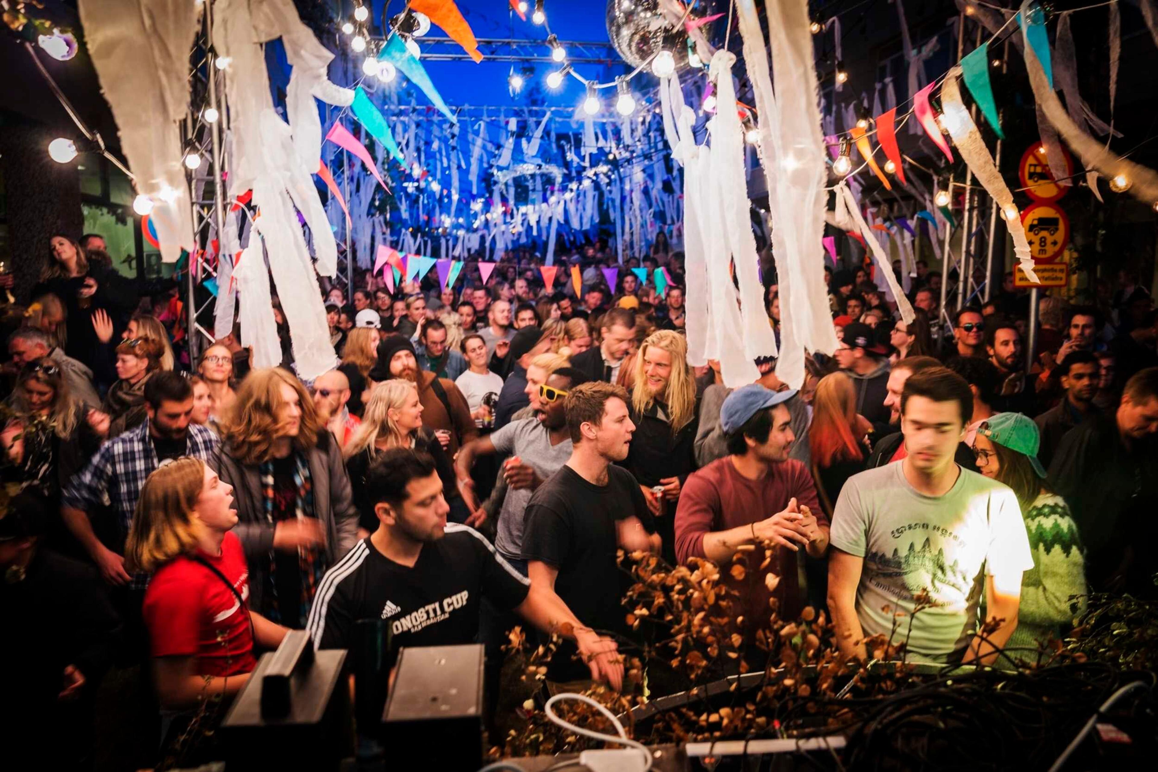 Crowded outdoor festival at night with colorful lights, streamers, and bunting, featuring people socializing and enjoying the lively atmosphere.