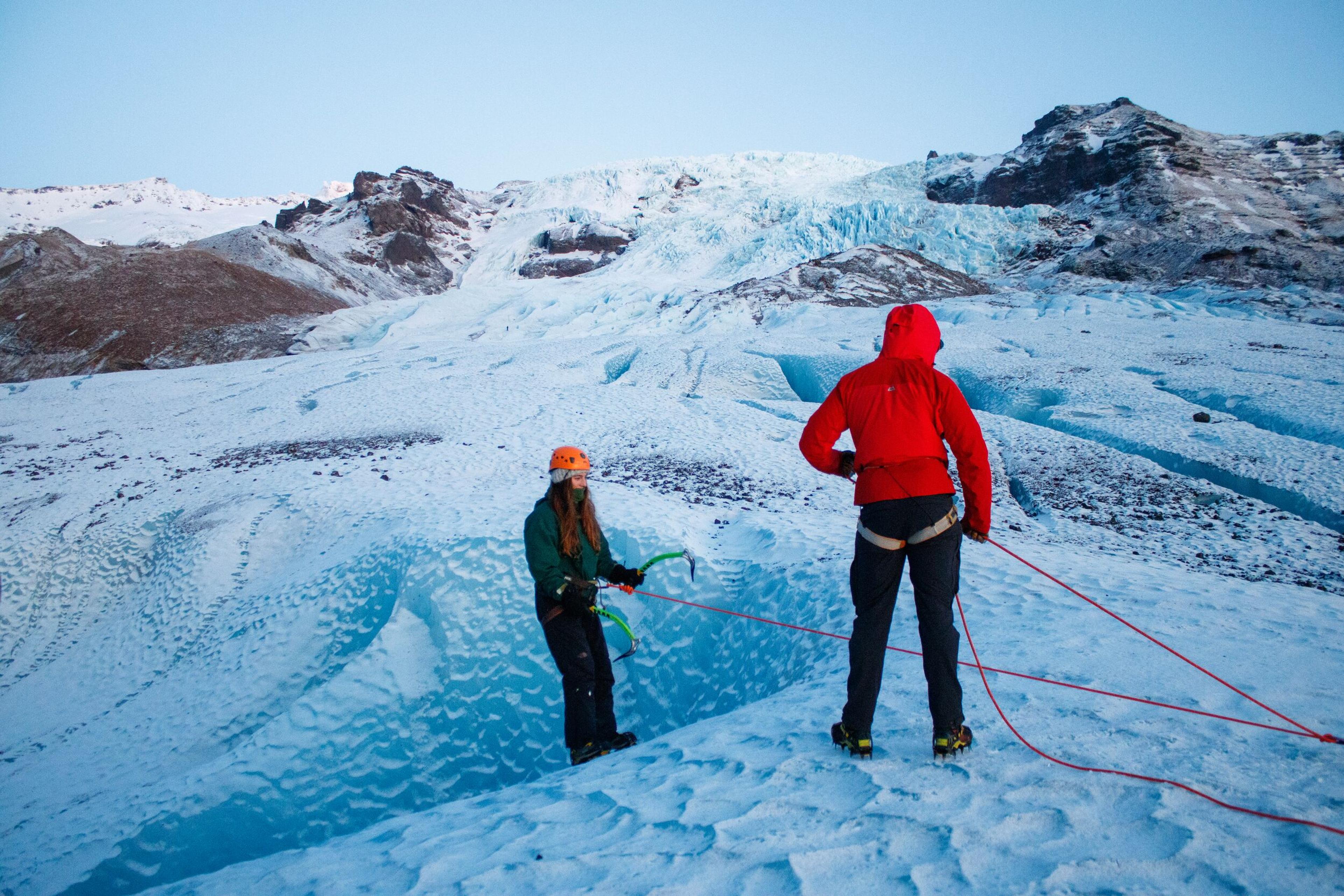 Two people on a glacier, one with an ice axe and the other handling ropes, with snow-covered mountains in the background.