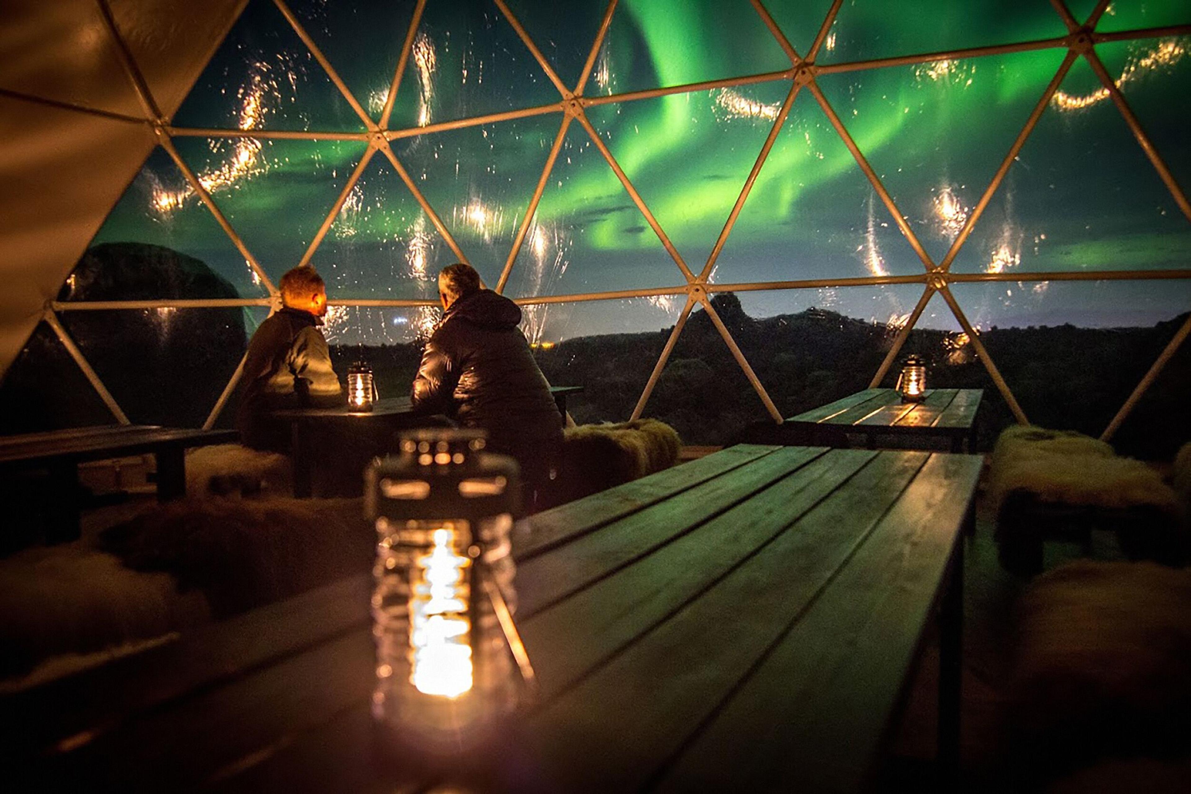 Interior of a geodesic dome at Aurora Base Camp, Iceland, with lanterns glowing and northern lights visible through the dome windows.