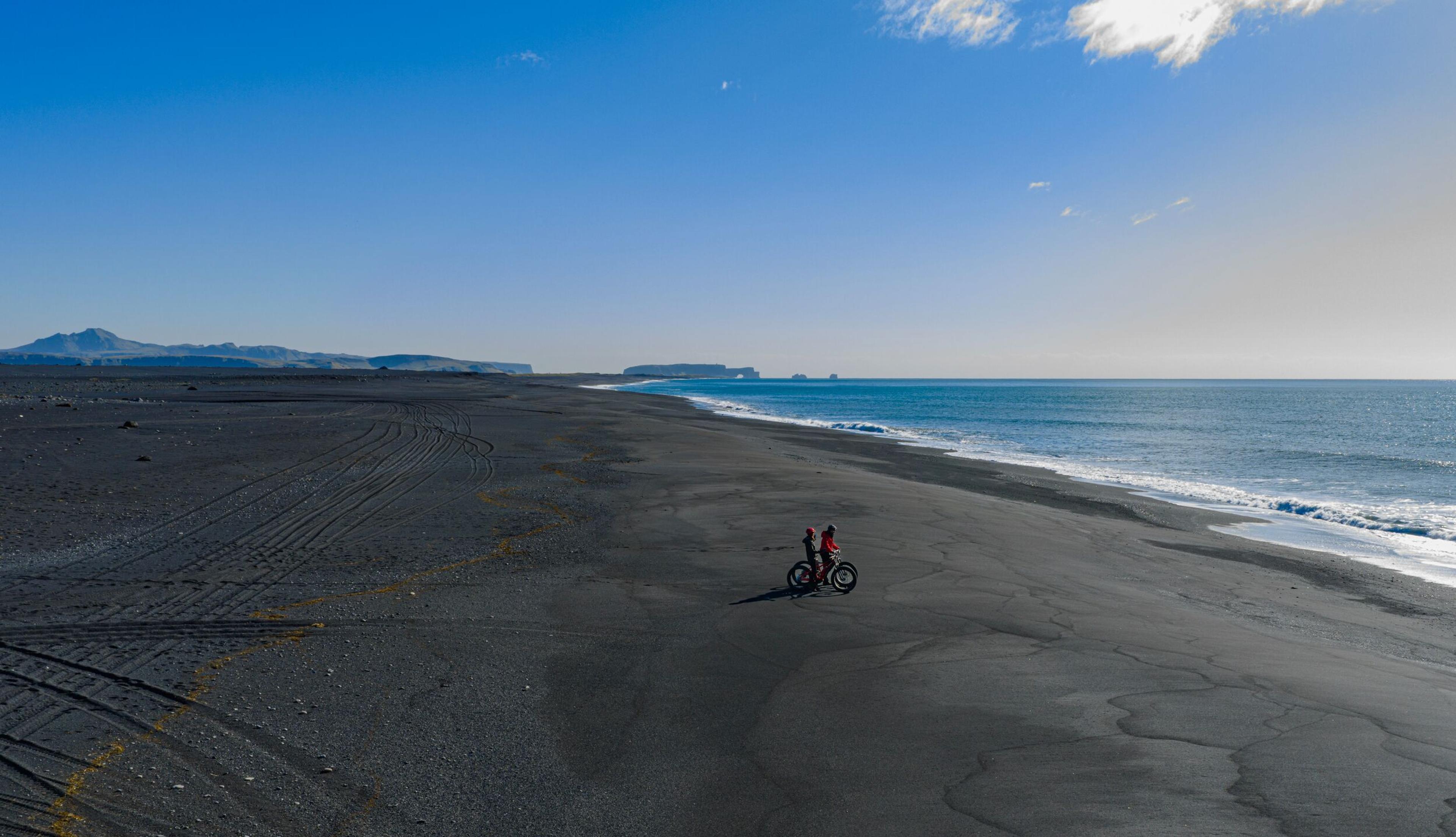 Two people with fat bikes on a black sand beach beside the ocean.