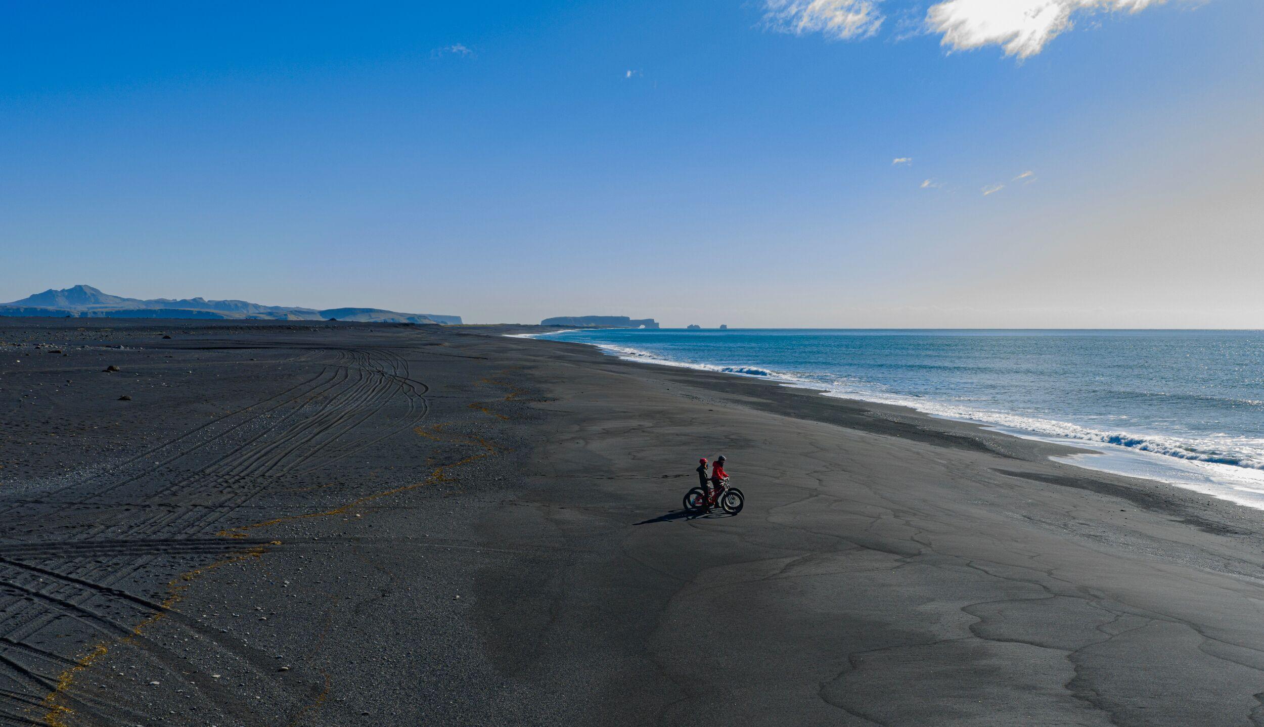 Two people with fat bikes on a black sand beach beside the ocean.