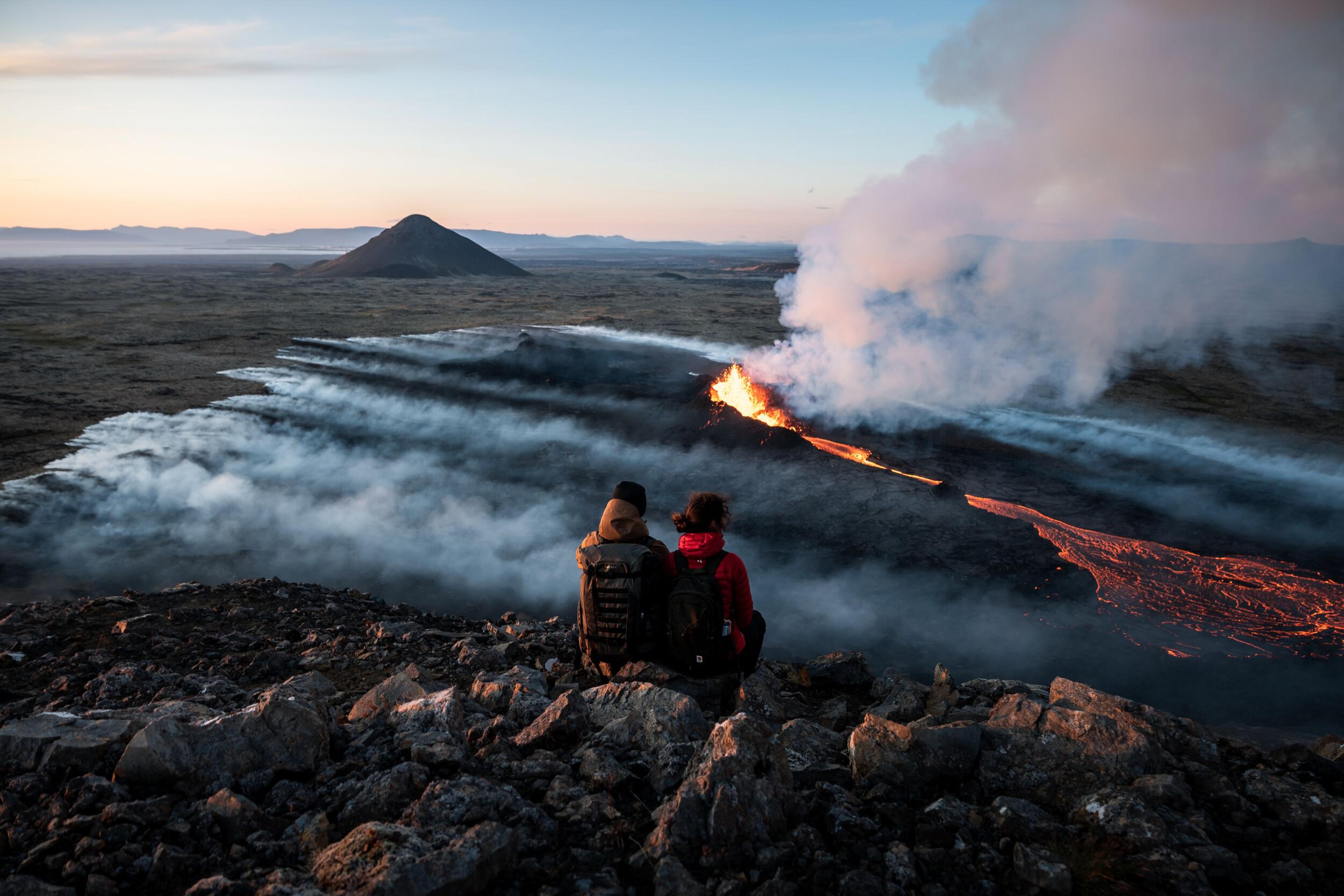 "Two people observing a dramatic volcanic eruption in Iceland, with lava flows and smoke.
