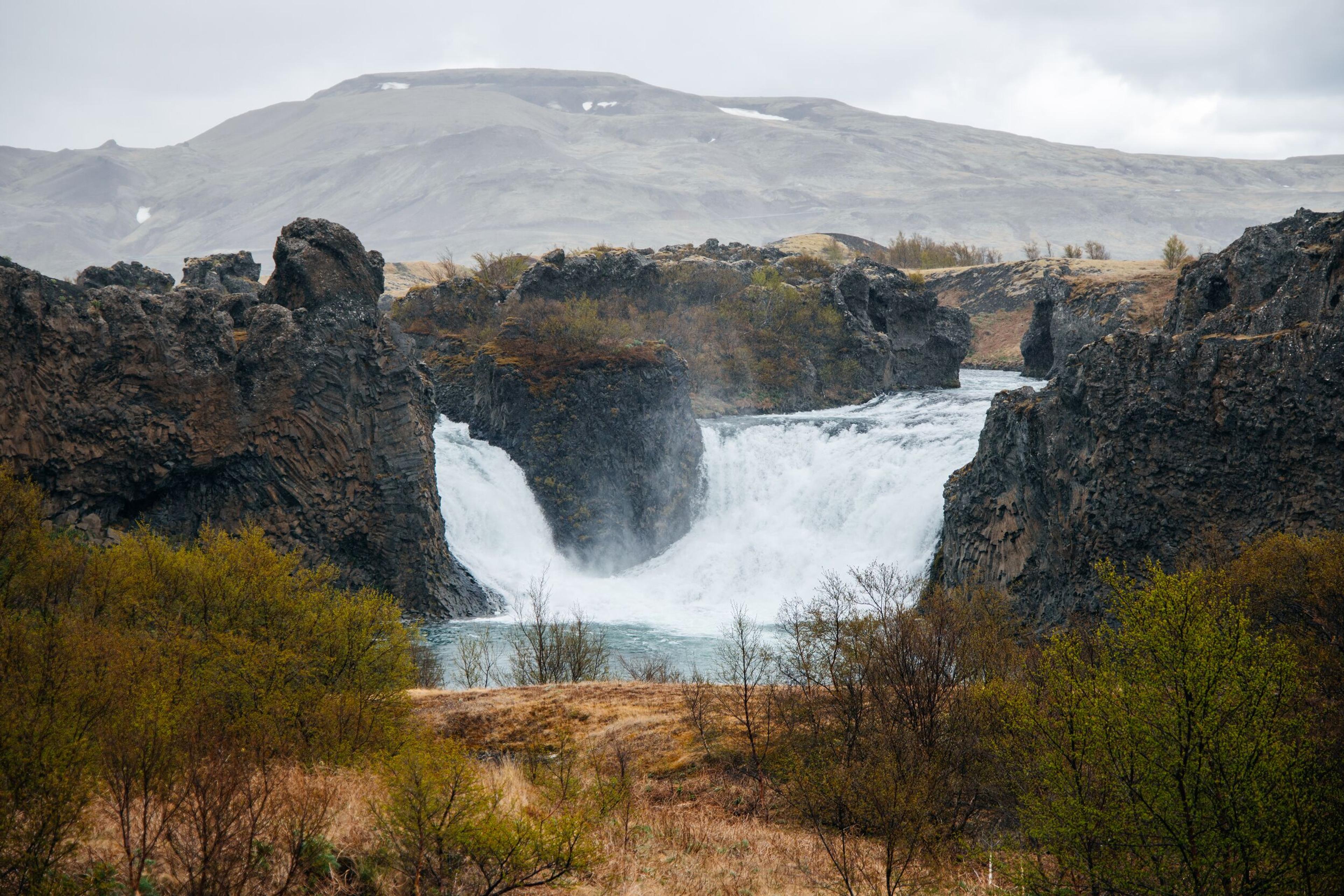 A powerful waterfall cascades between dark, rugged cliffs under a cloudy sky.