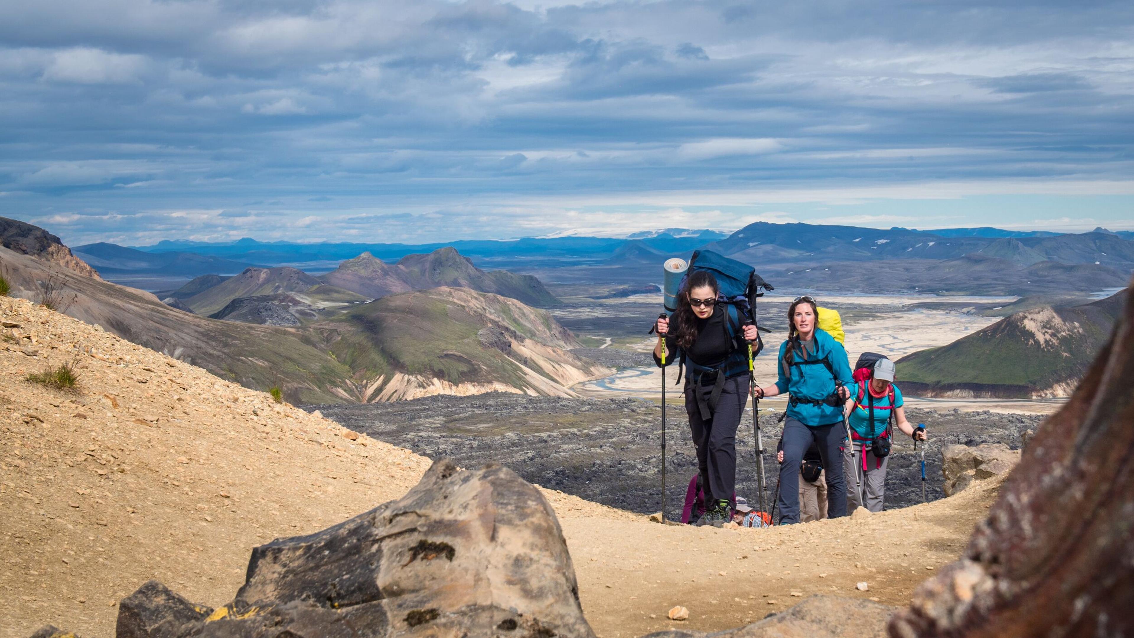 Three hikers with backpacks and poles climb a sandy, rocky mountain trail, with a sweeping view of distant mountains and valleys under a cloudy sky.
