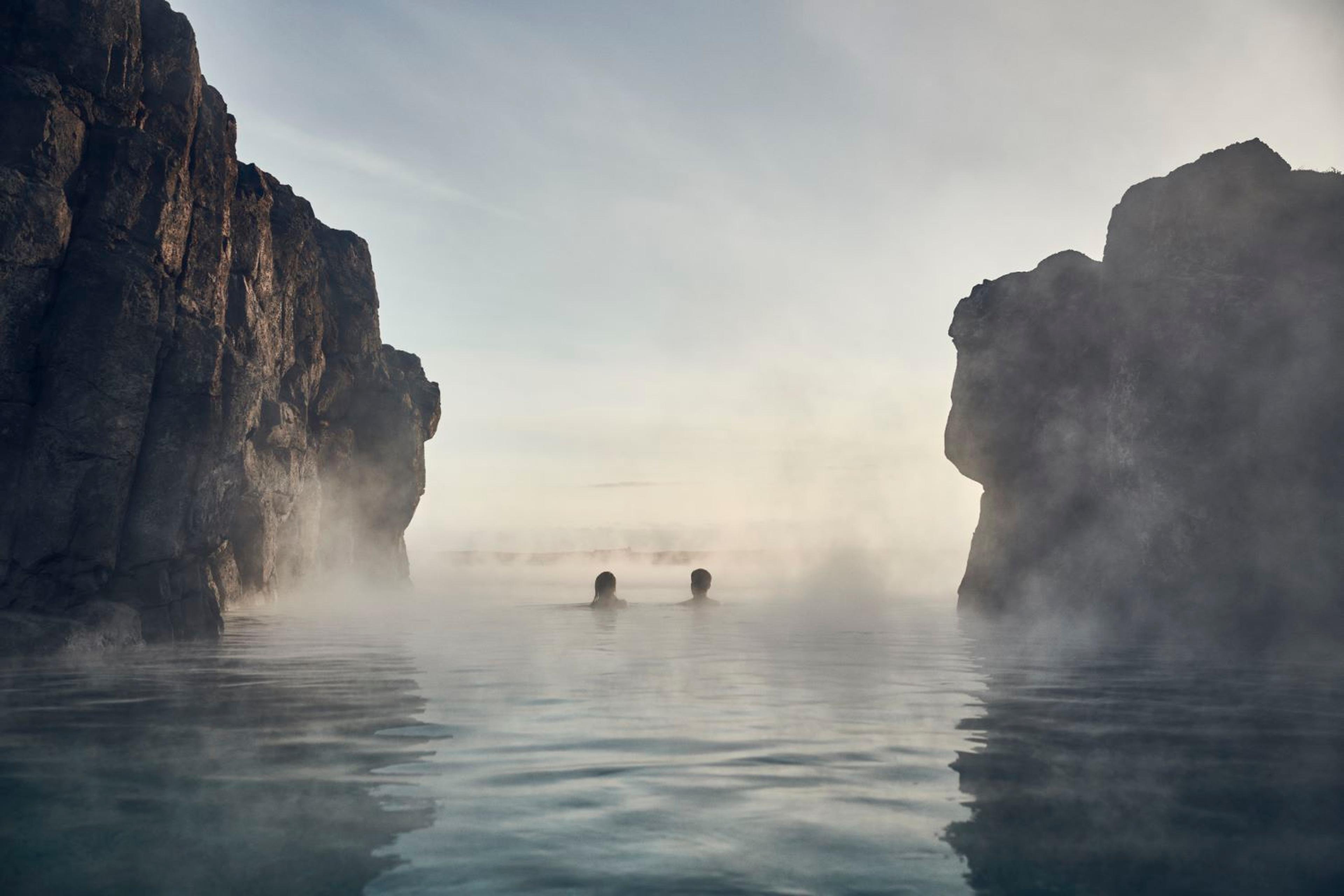 Two people bathe in a steamy, blue geothermal pool between dark cliffs.